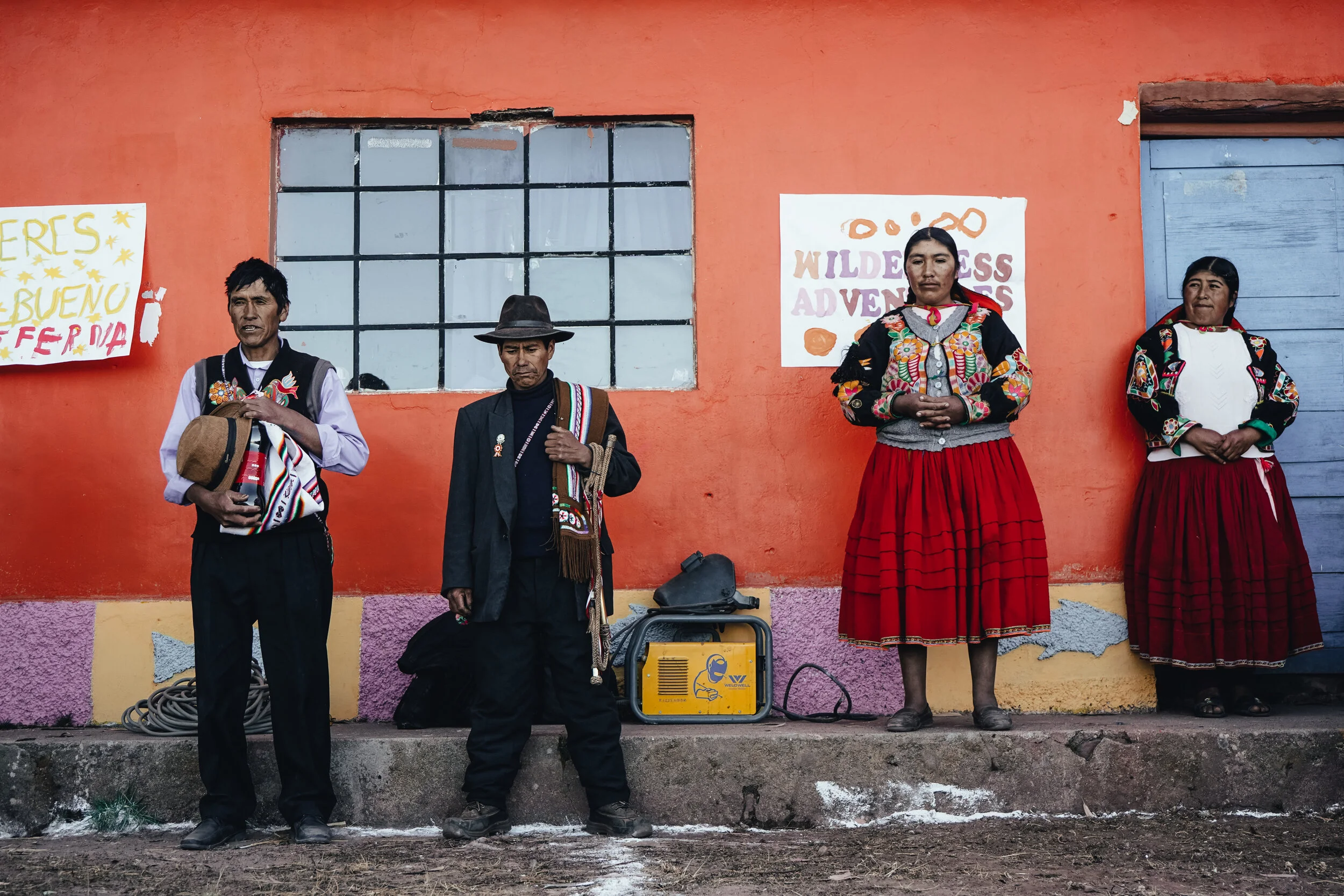 Four indigenous people standing in front of a painted orange wall, two women on the right wearing traditional embroidered blouses and red skirts, two men on the left wearing traditional attire, one holding a hat. There are signs on the wall reading "