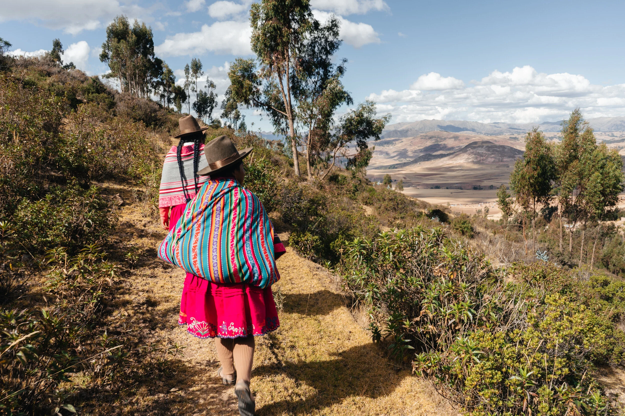 Two women wearing colorful traditional clothing and hats walk along a dirt trail in a hilly landscape with trees and distant mountains under a partly cloudy sky.