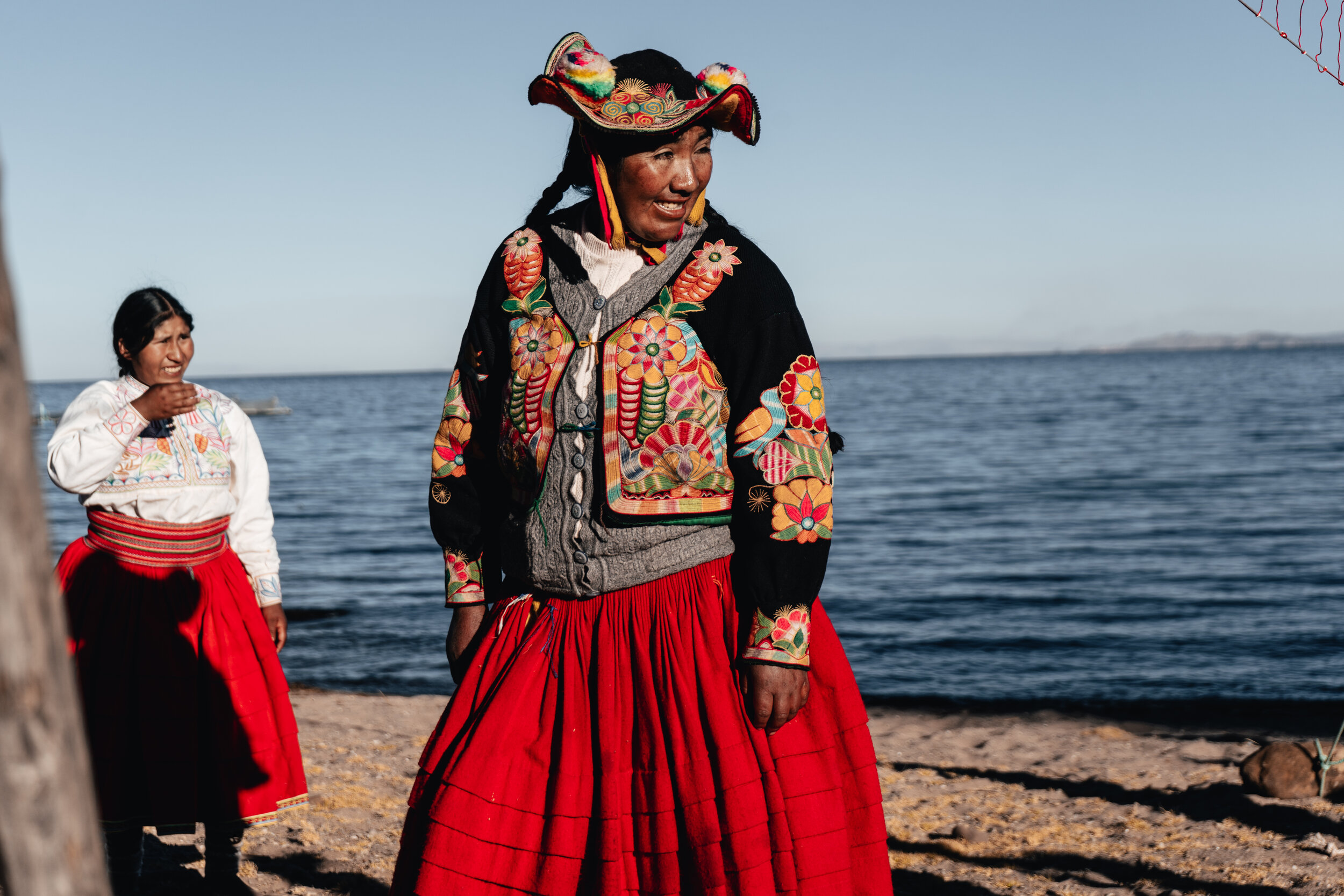 Two women in colorful traditional Mexican clothing standing on a beach with water in the background. One woman is in the foreground wearing a embroidered jacket, a gray sweater, a wide-brimmed hat, and a long red skirt. The other woman is in the back