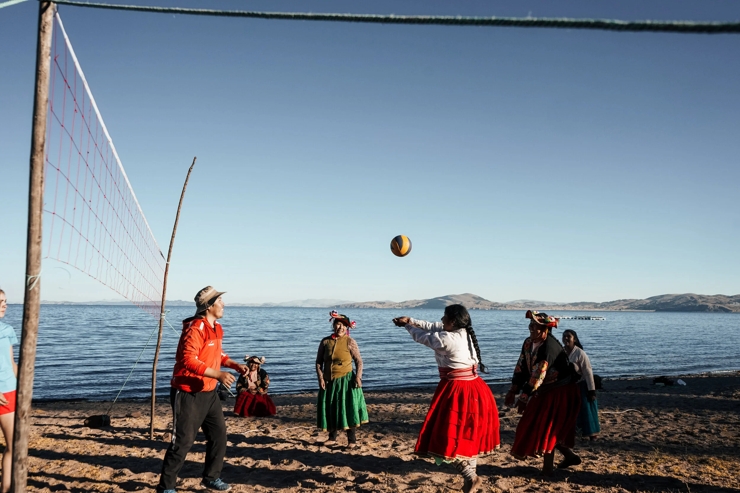 People playing beach volleyball near a body of water, with a sandy shore and distant hills under a clear sky.