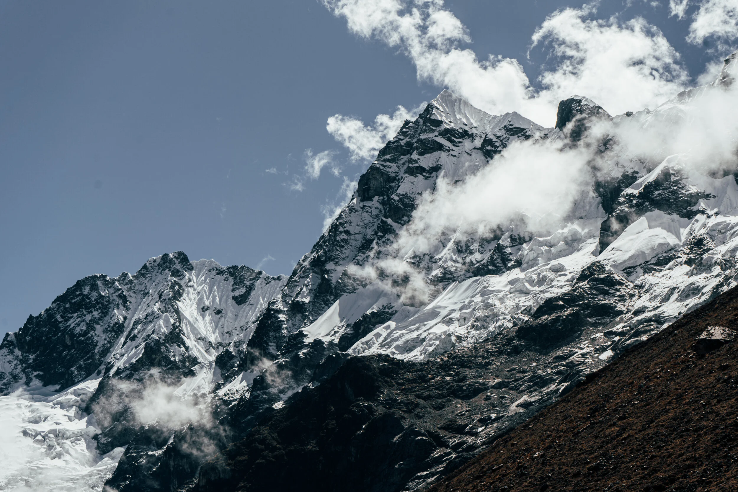 Snow-capped mountain peaks with ice, icefall and snow, rugged dark rocks on lower slopes, against a partly cloudy sky.