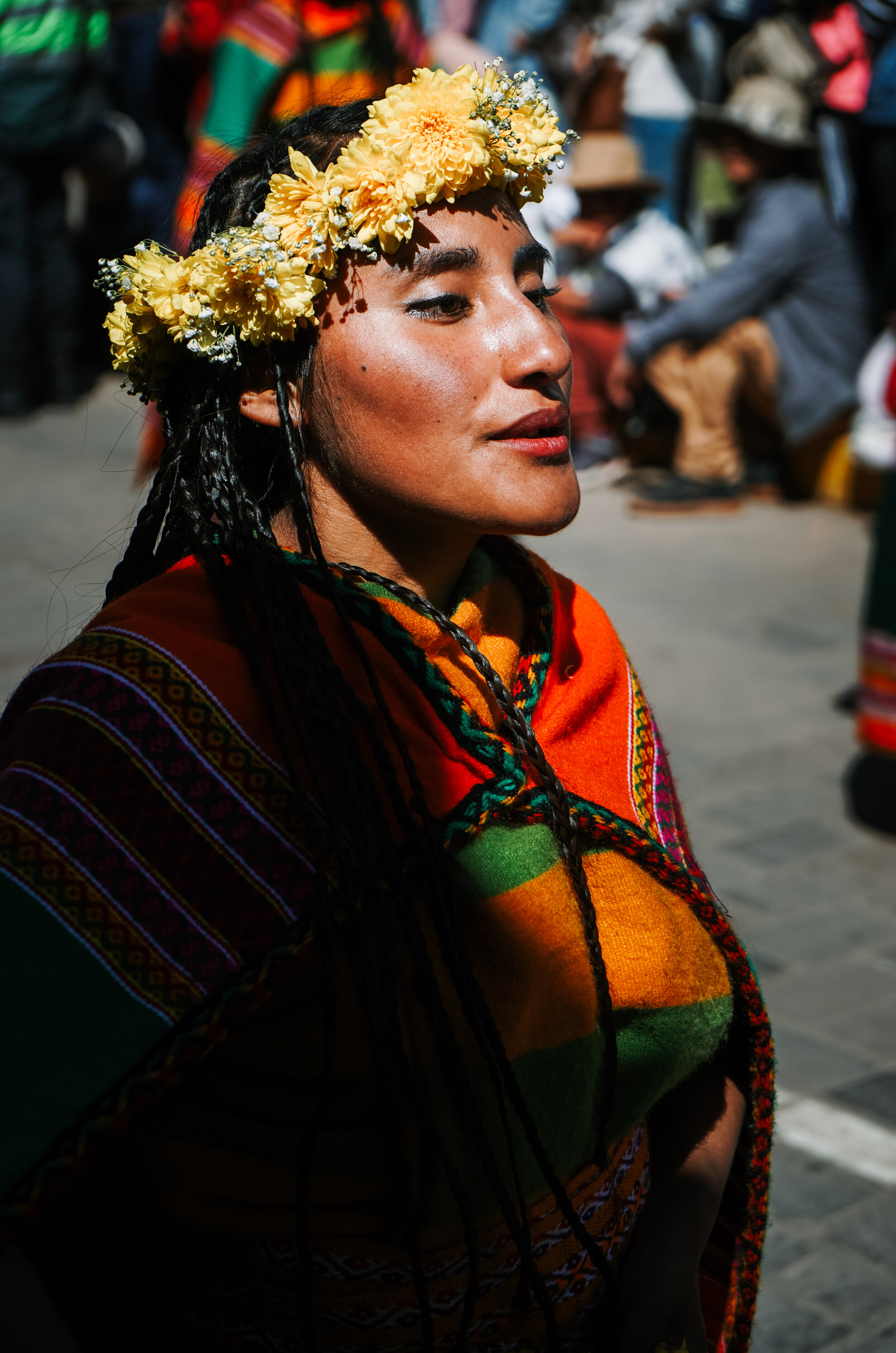 A woman with dark braided hair wearing a yellow flower crown, colorful traditional clothing, and red lipstick, standing outdoors during a cultural event or celebration.