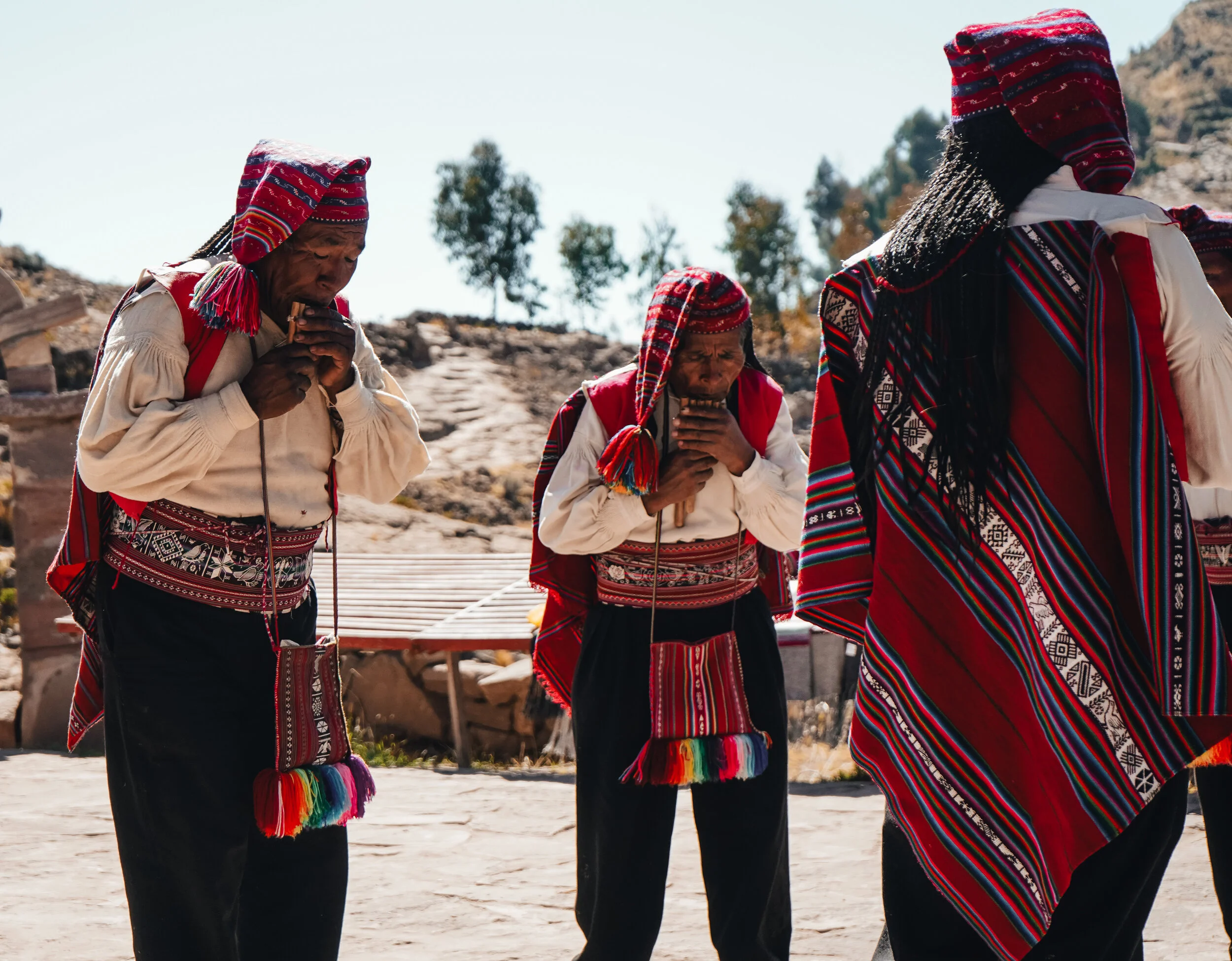 Three indigenous people dressed in traditional colorful clothing participating in a ceremony outdoors.