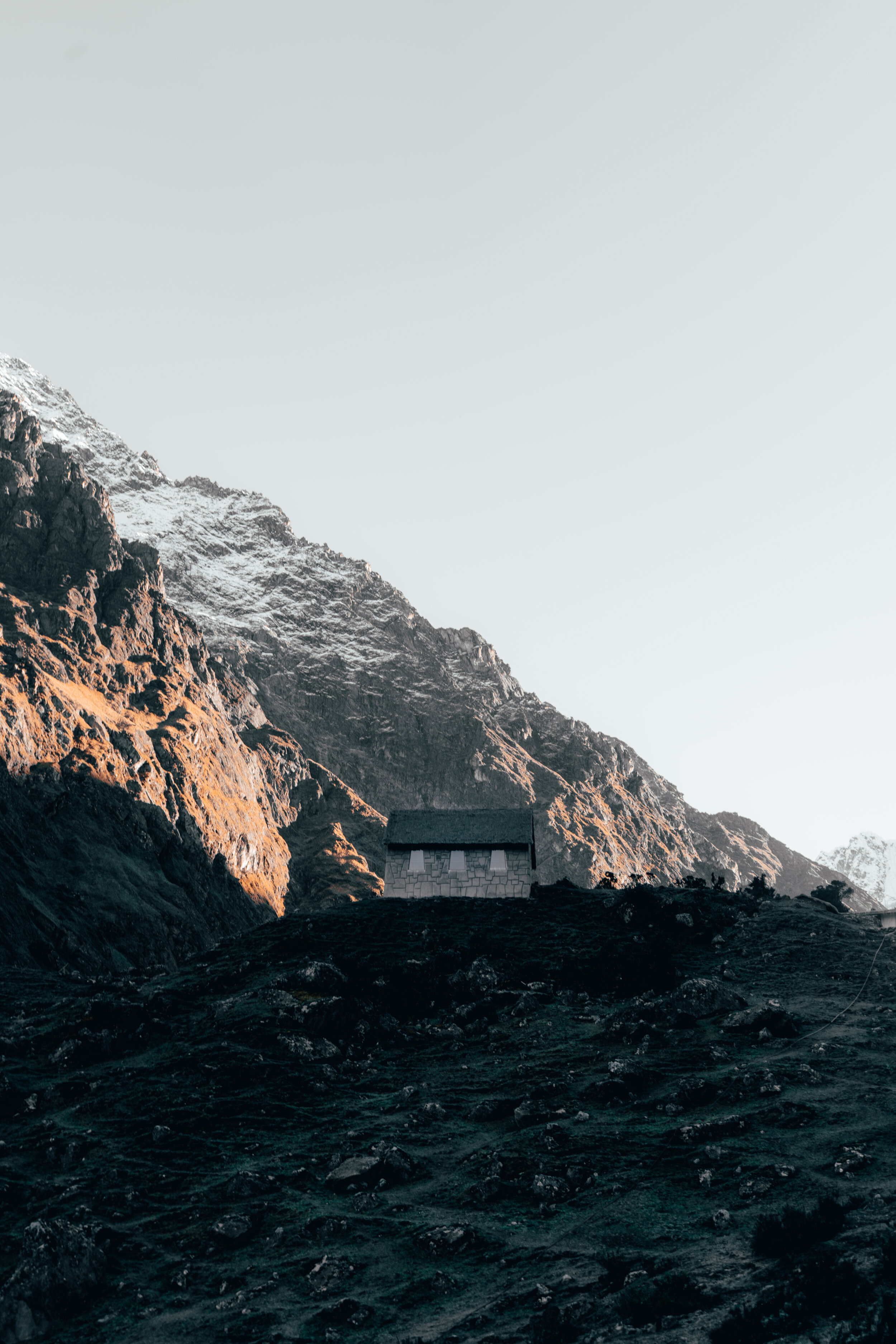 A small house sits on dark, rugged terrain at the base of a snow-capped mountain range, with a clear sky overhead.