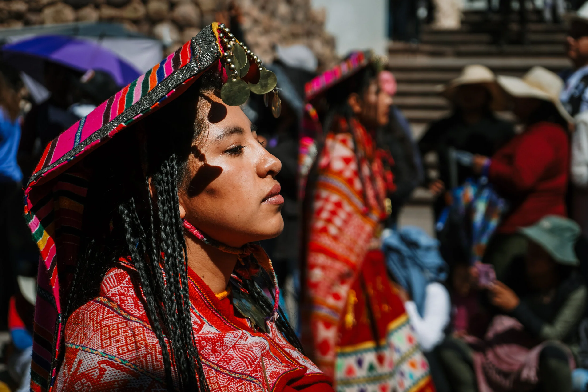 A woman in traditional colorful clothing and a decorated hat with coins, standing in a crowd with similar attire and hats, outdoors in sunlight.