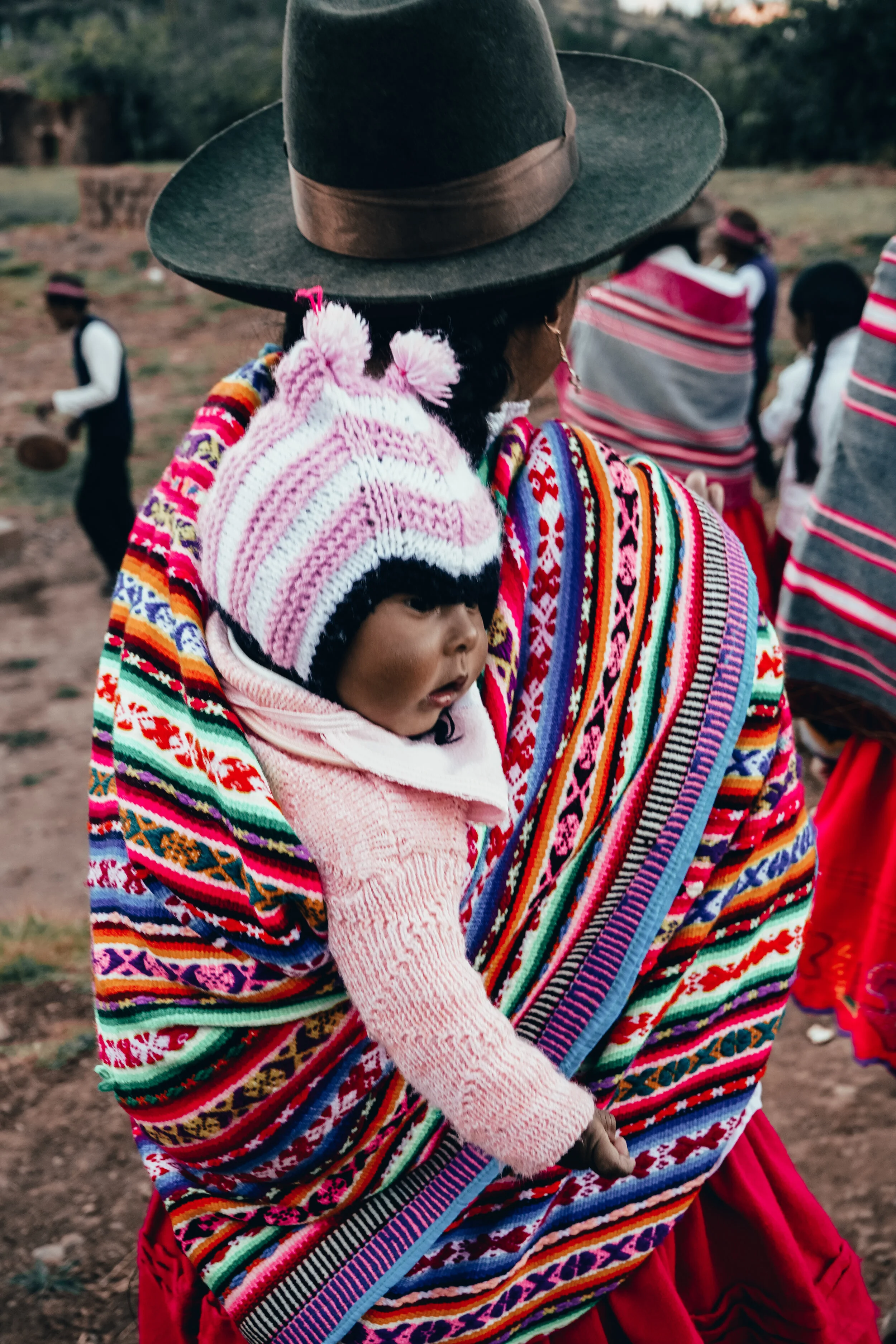A woman carrying a child on her back, both dressed in traditional colorful clothing, during an outdoor cultural event or celebration. In the background, other people are visible, also dressed in traditional attire.