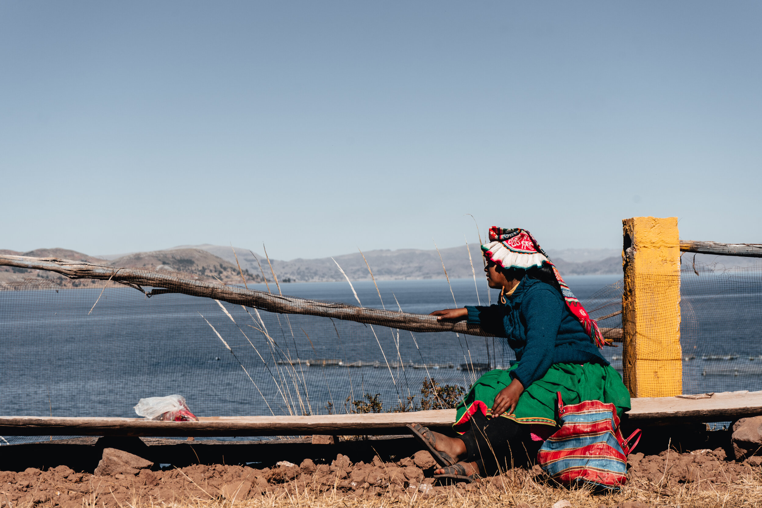 A woman dressed in traditional clothing, sitting on the ground near a wooden fence with a body of water and hills in the background.