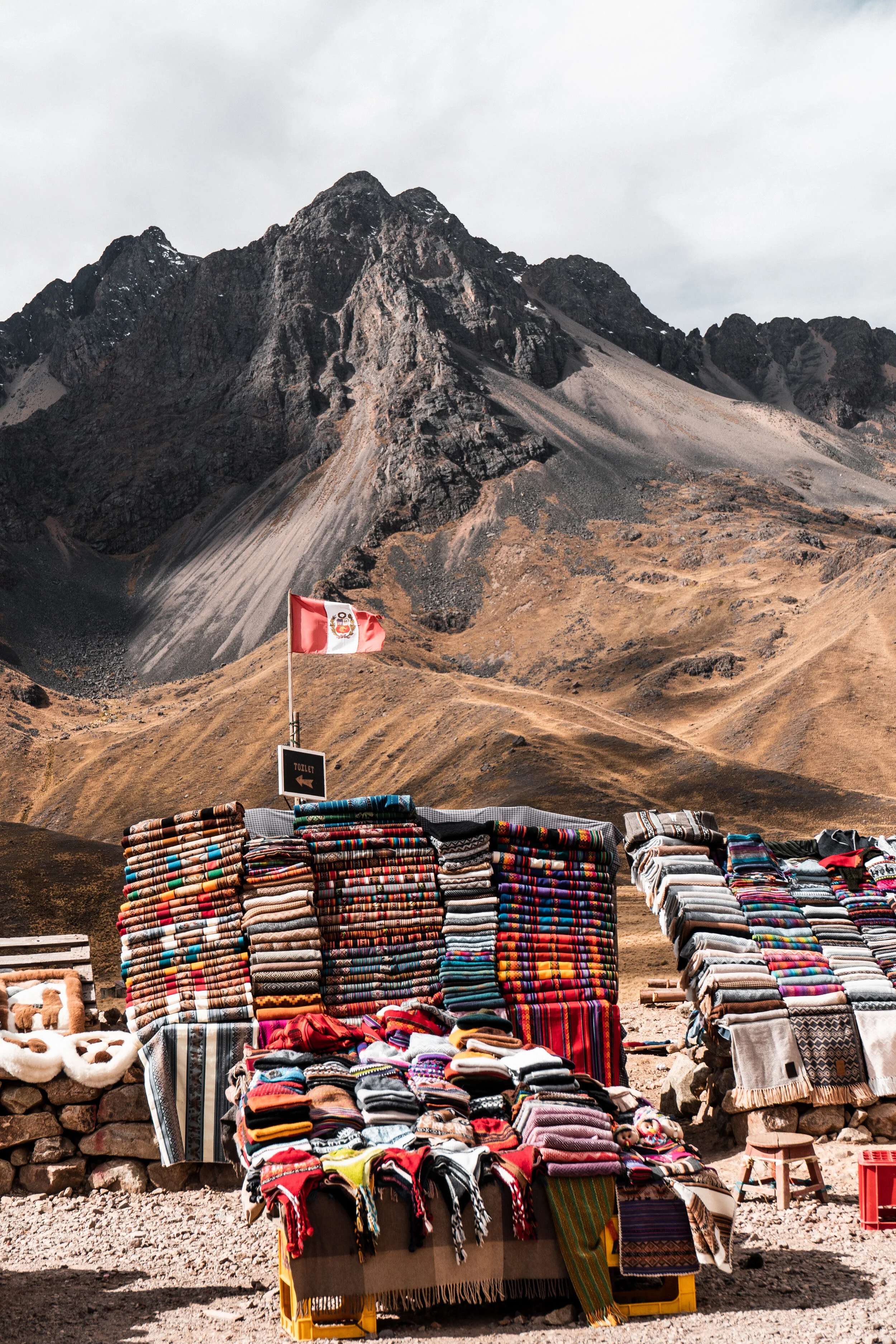 An outdoor market stall displaying colorful woven blankets and textiles against a mountainous landscape with a Peruvian flag.
