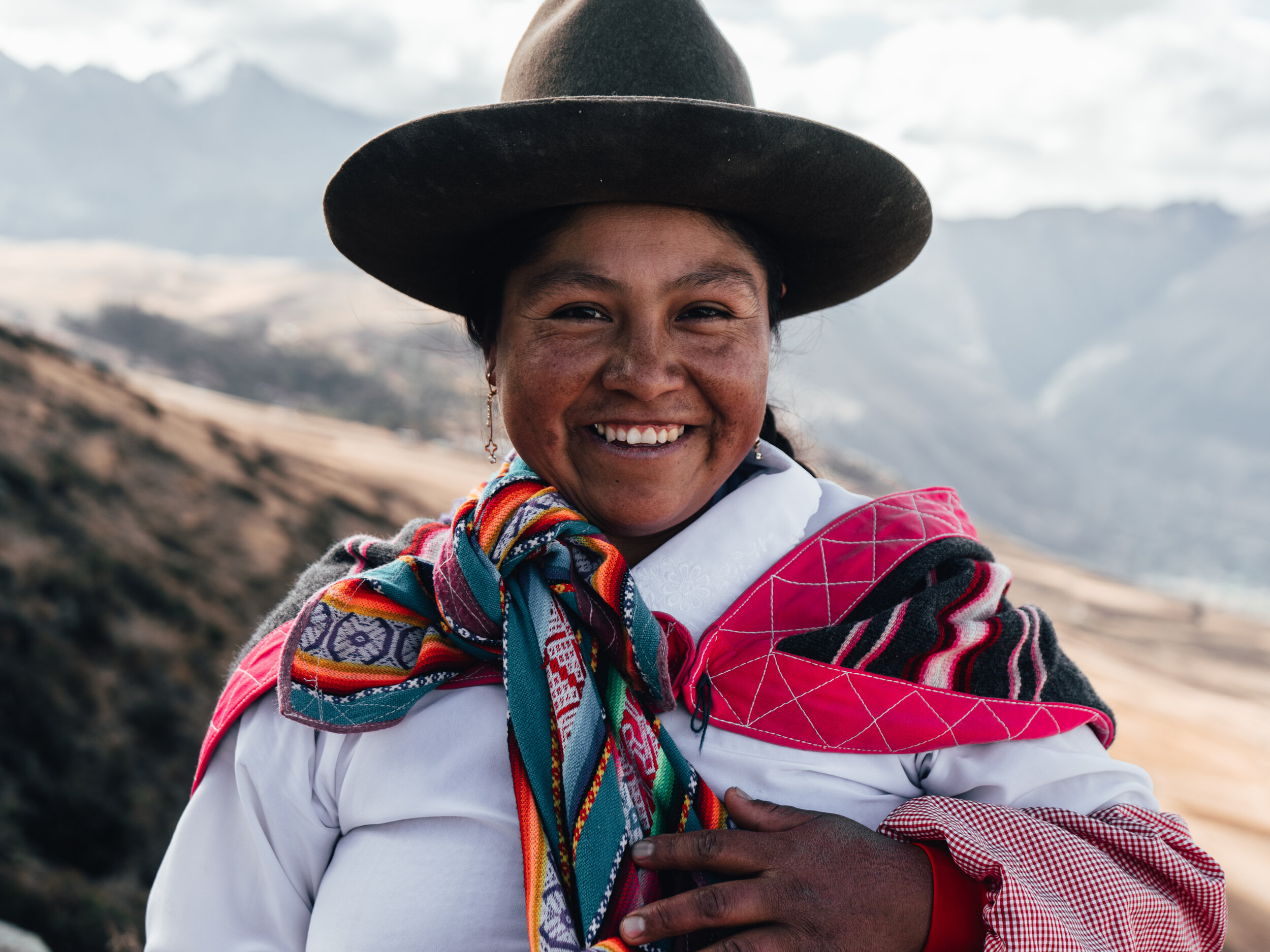 A smiling woman dressed in traditional Andean clothing, wearing a wide-brimmed hat and a colorful scarf, standing outdoors with mountain scenery in the background.