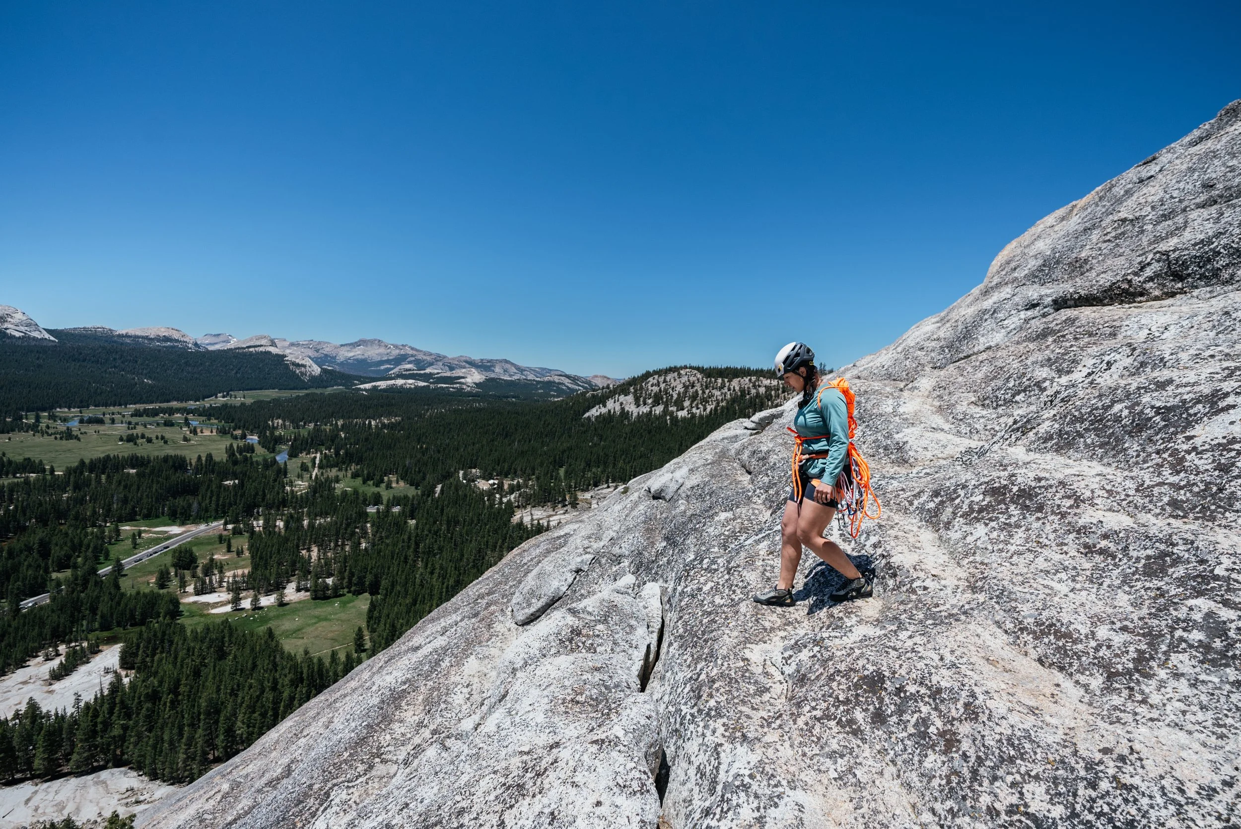A person wearing a helmet and climbing gear is ascending a steep granite rock face, overlooking a broad forested valley and mountain range under a clear blue sky.