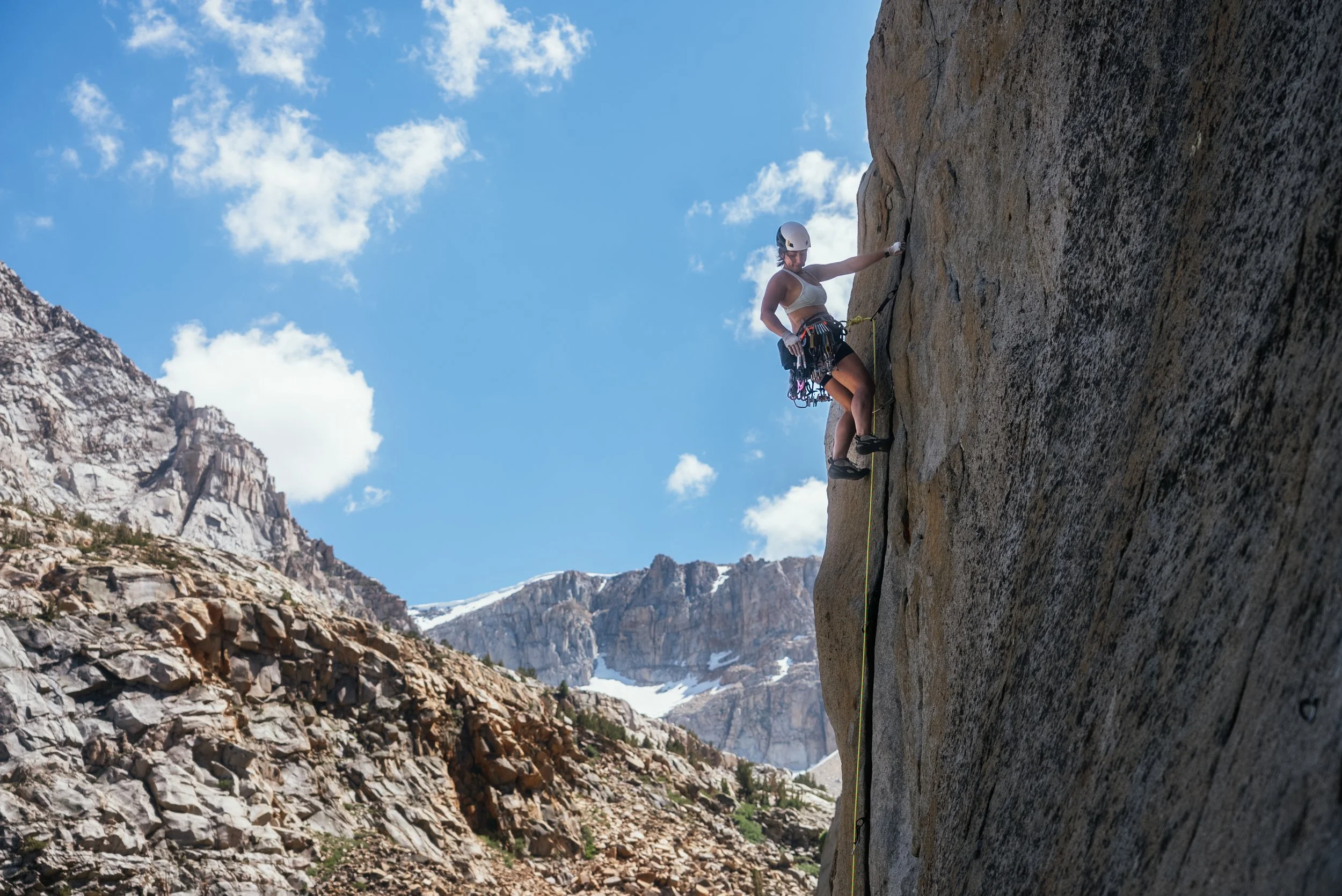 A woman rock climbing on a steep cliffside with a mountain landscape and blue sky with clouds in the background.