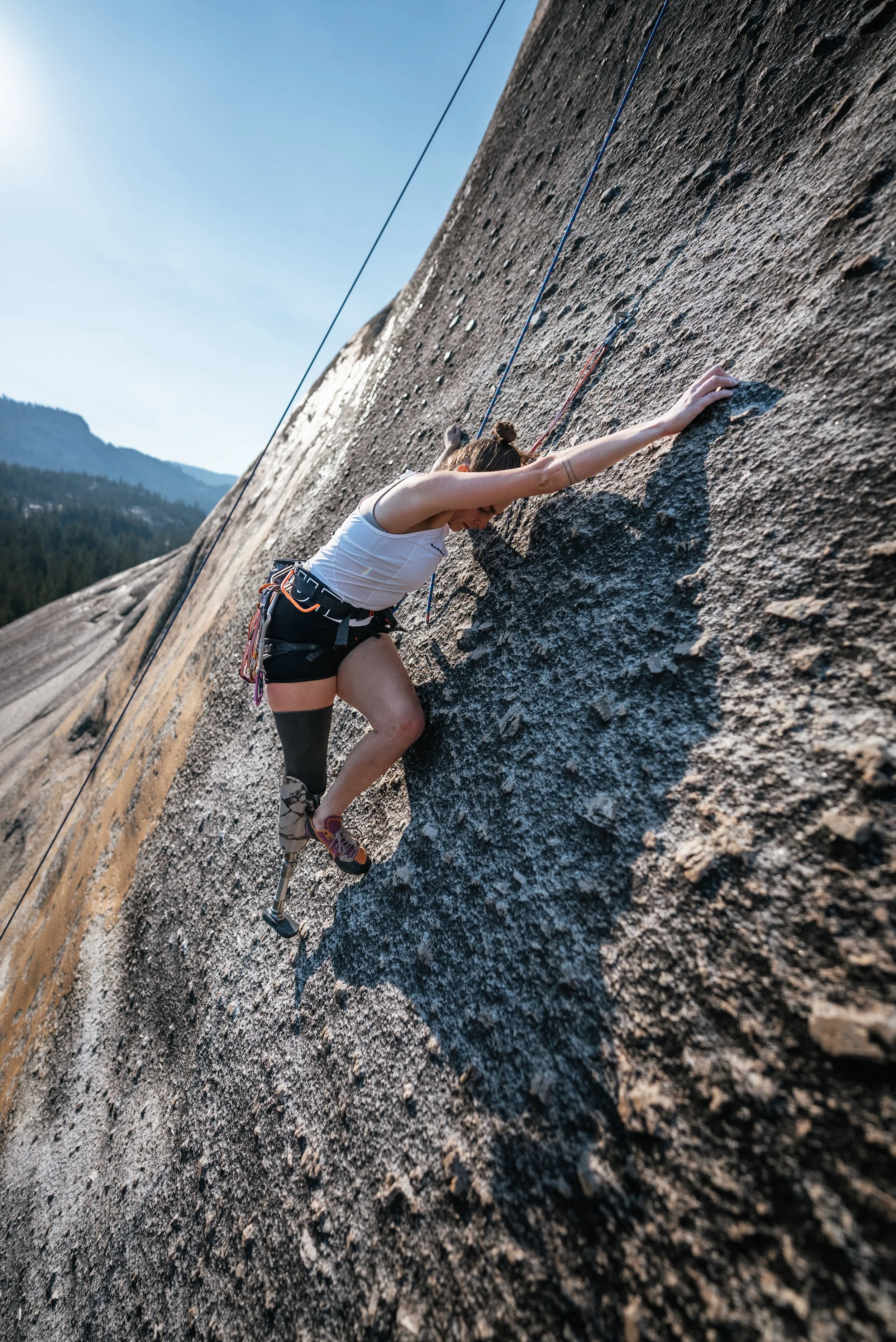 A woman rock climbing on a steep mountain wall with mountains and trees in the background.