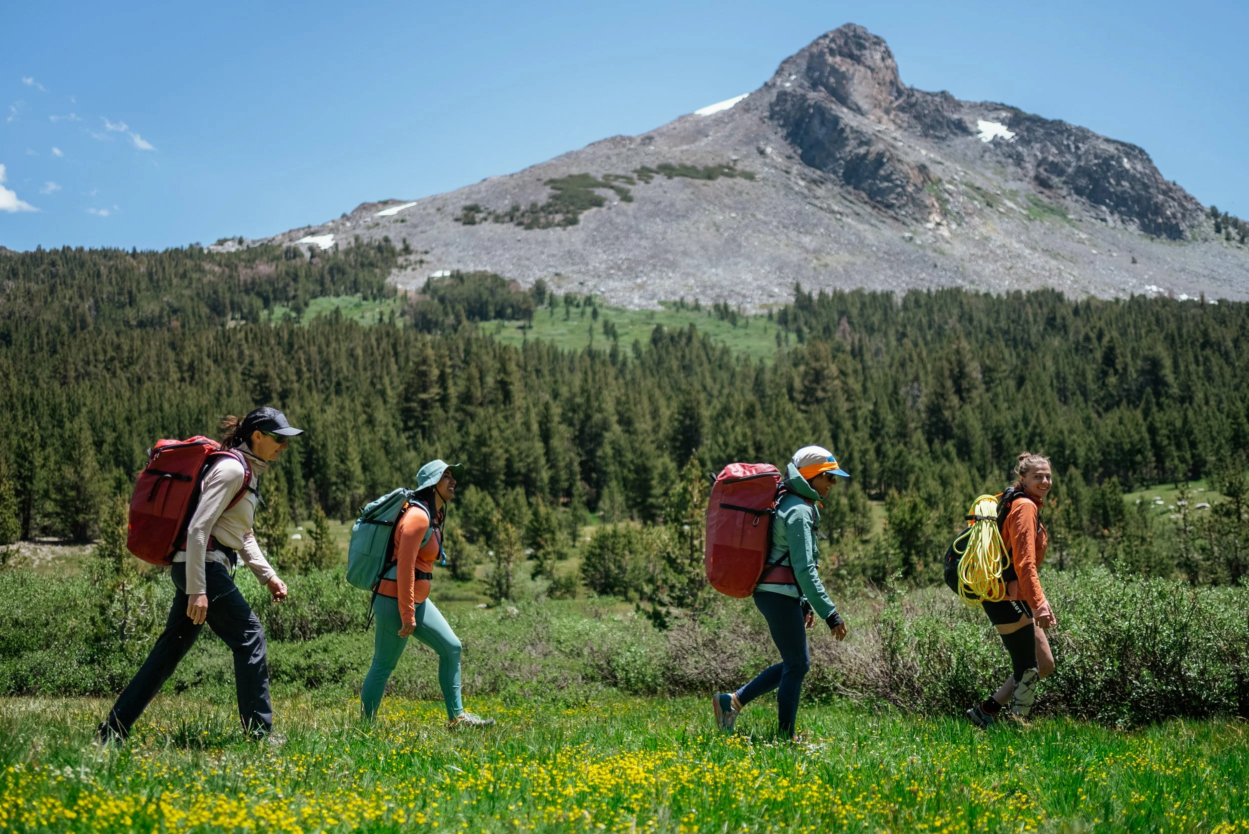 Four women hiking through a green field with trees and a mountain in the background on a sunny day.