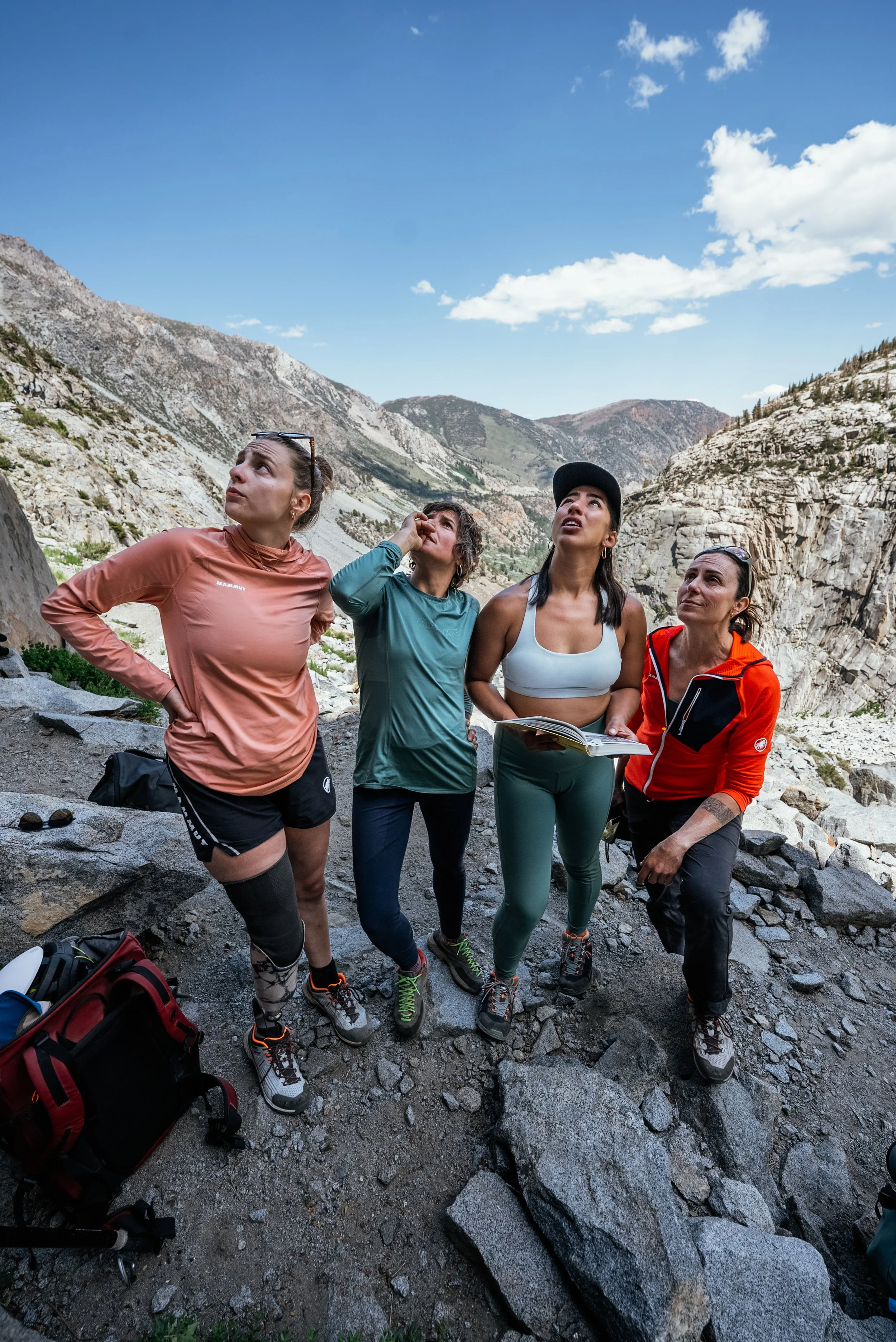 Four women hikers in athletic clothing standing on rocky terrain in a mountainous area, looking up and pointing at something in the sky on a clear day.