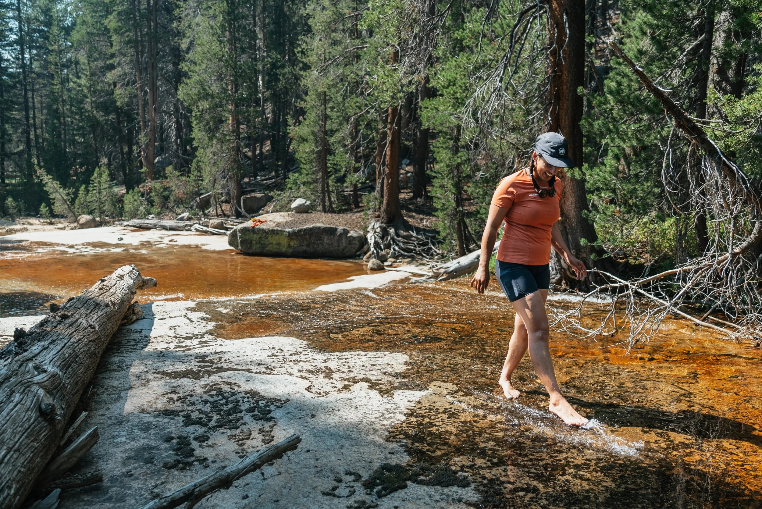 A woman in an orange T-shirt and black shorts walking barefoot across a shallow, rocky stream in a wooded forest.