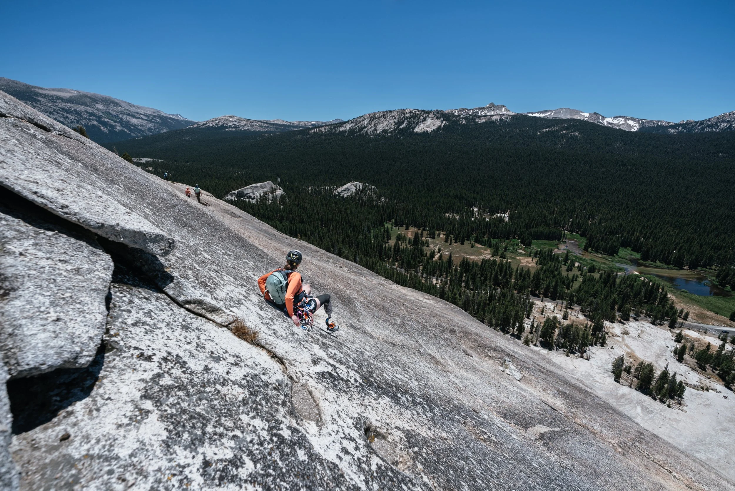 A person with a helmet and backpack climbing a large, sloped granite rock formation in a mountainous landscape with forested valleys and distant snow-capped mountains under a clear blue sky.