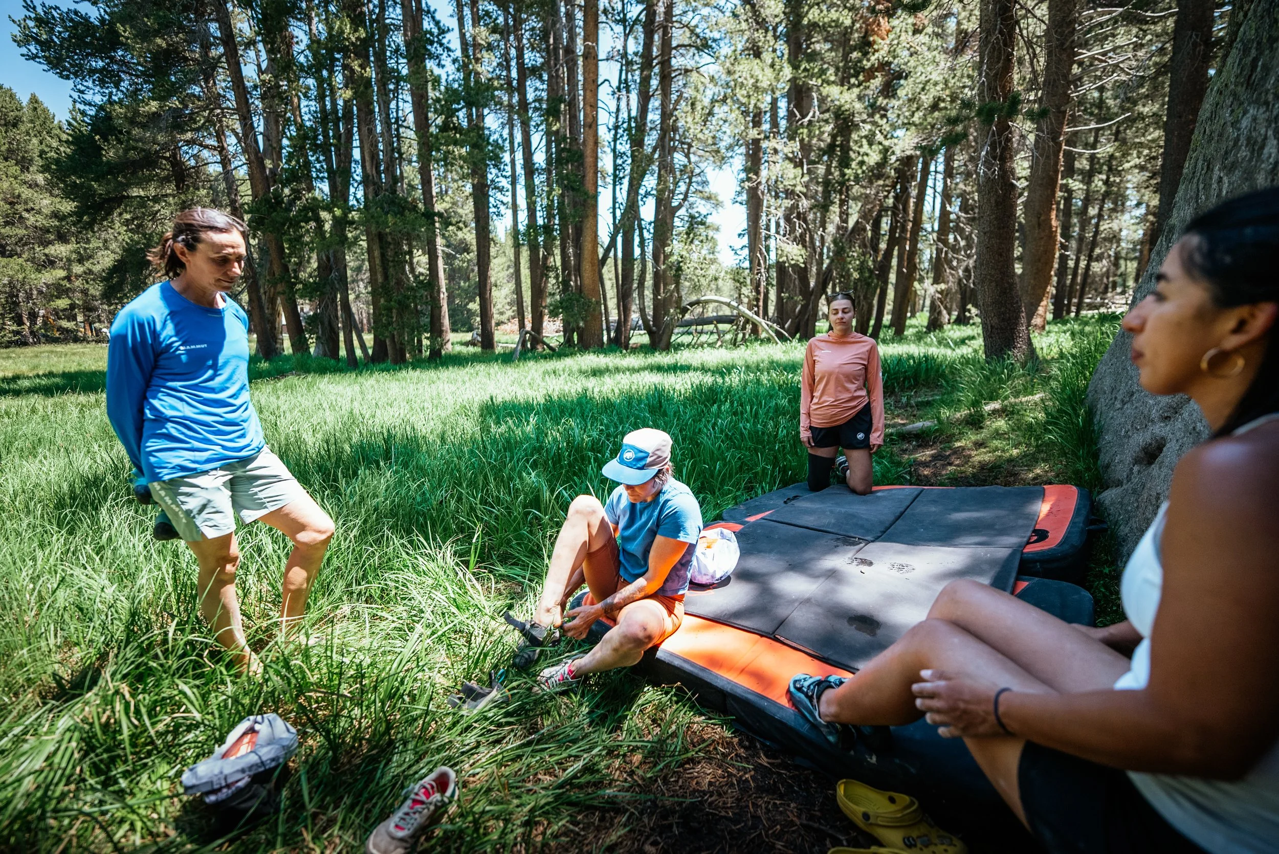Group of women in a forest clearing, one sitting on the ground tying her shoe, others seated or standing nearby with a padded mountain rescue mattress, surrounded by tall trees and green grass.