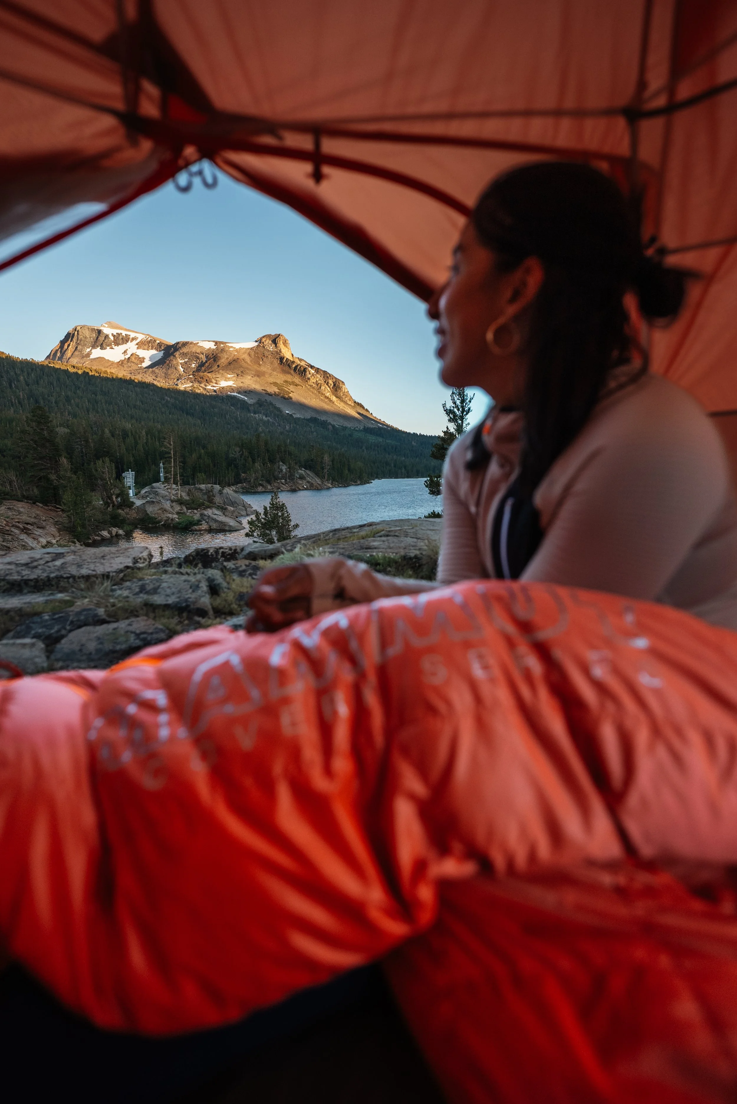 A woman sitting under a camping tent, overlooking a lake and mountain landscape with snow-capped peaks and pine trees, during sunset.