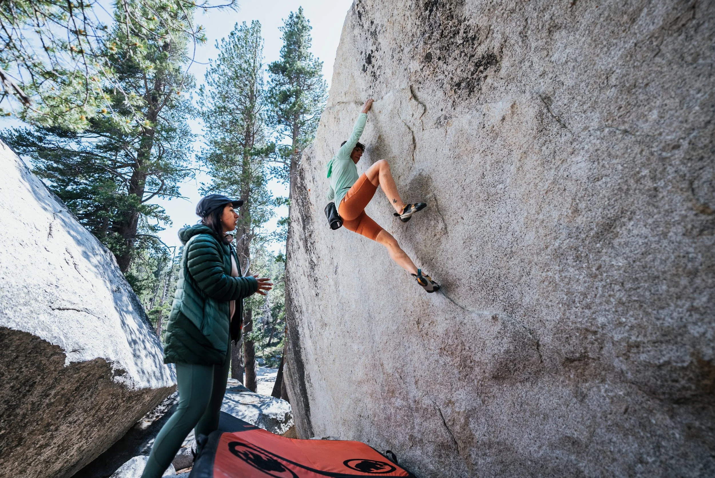 A woman wearing climbing gear, including climbing shoes and chalk, is rock climbing on a large, textured indoor climbing wall outdoors, with a woman instructor observing and giving advice nearby.