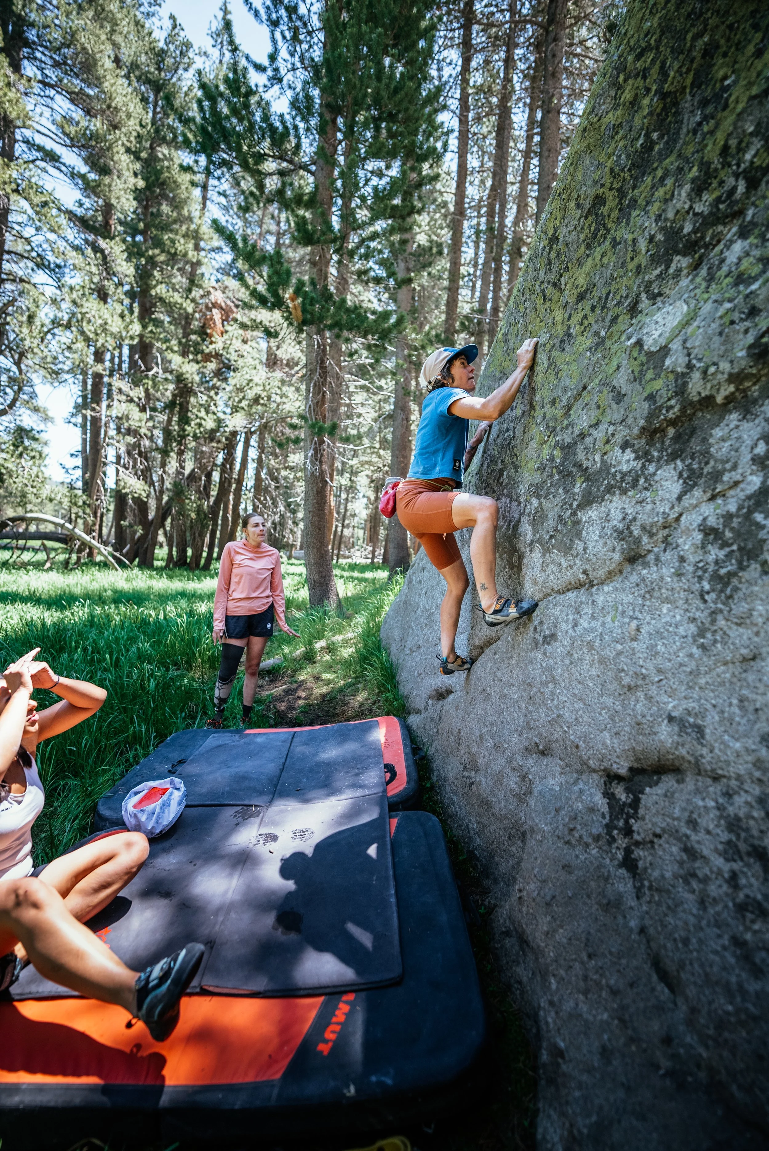 A woman rock climbing outdoors on a large boulder with the help of a crash pad below, while another woman observes in a forested area.