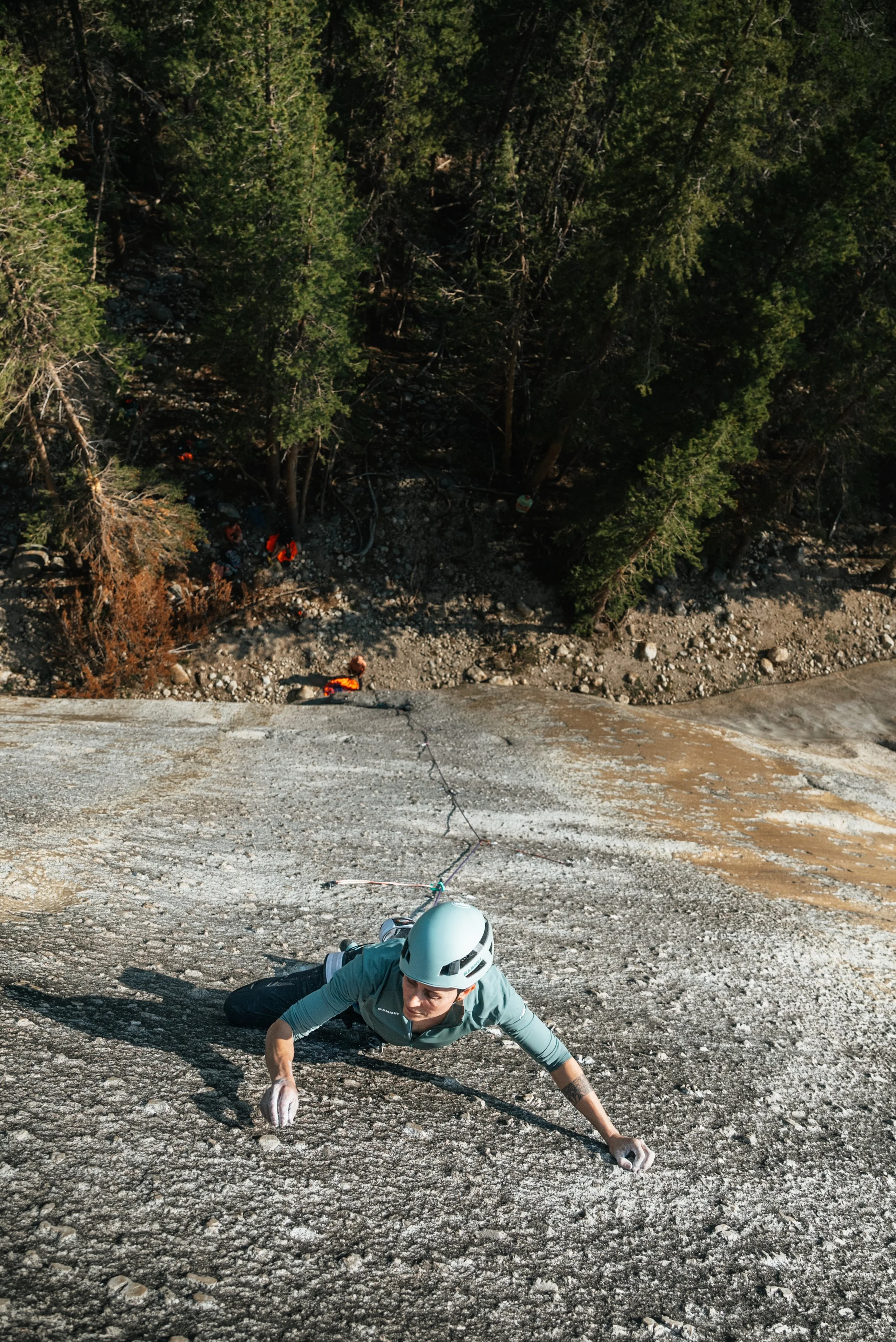 A person wearing a helmet and climbing gear is rock climbing on a steep granite surface, with a forested area below.