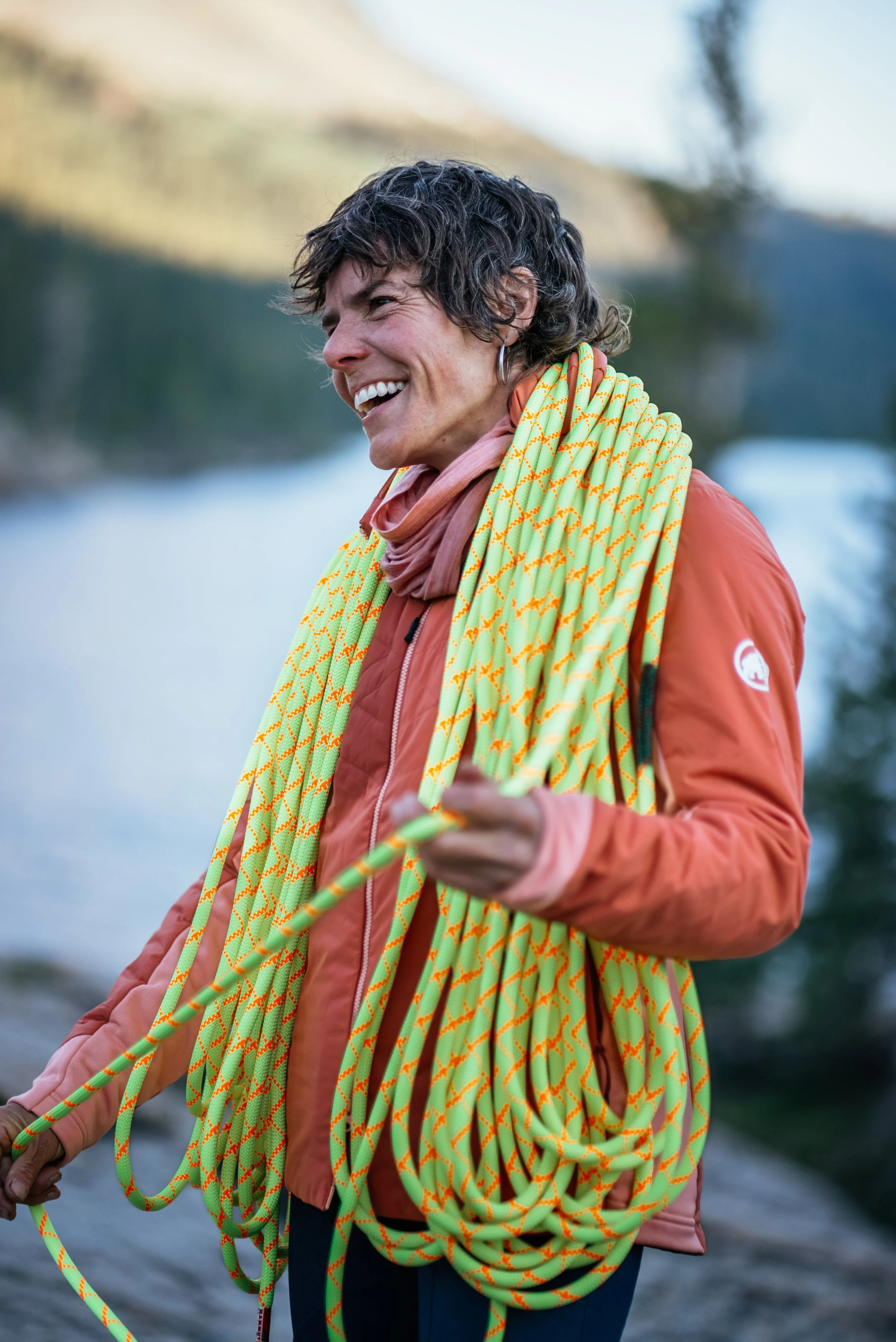A woman outdoors near a body of water, smiling, carrying a coiled climbing rope, wearing a coral jacket and a pink neck gaiter.