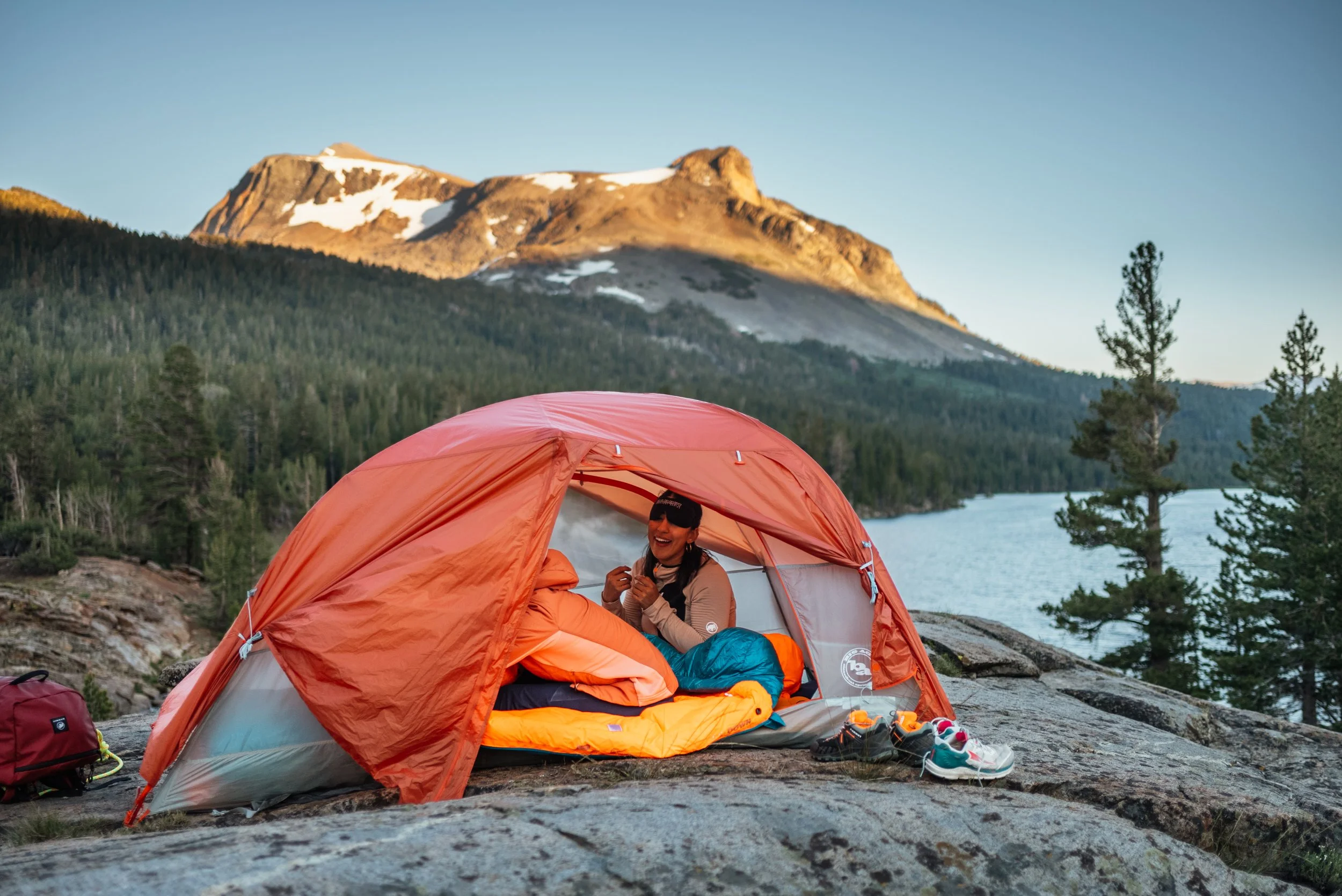A woman sitting inside a red tent on a rocky area near a lake, surrounded by trees and mountains, with a clear sky overhead.