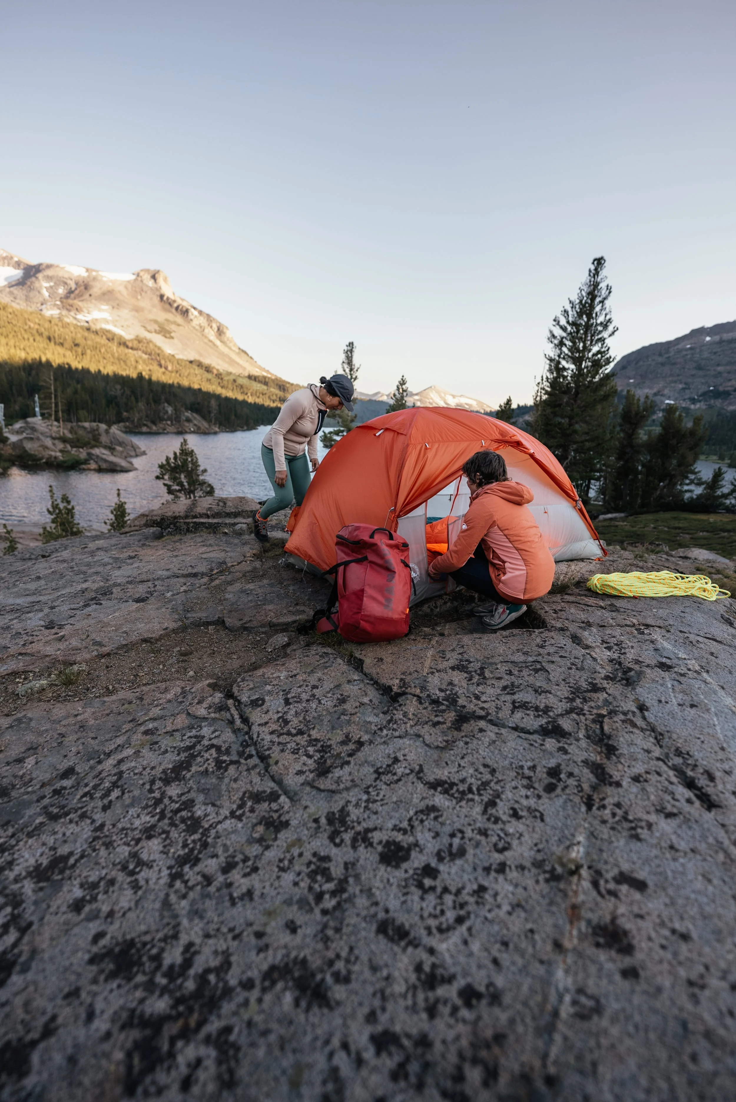 Two people setting up an orange camping tent on rocky ground beside a lake, with mountains and trees in the background, during sunset.