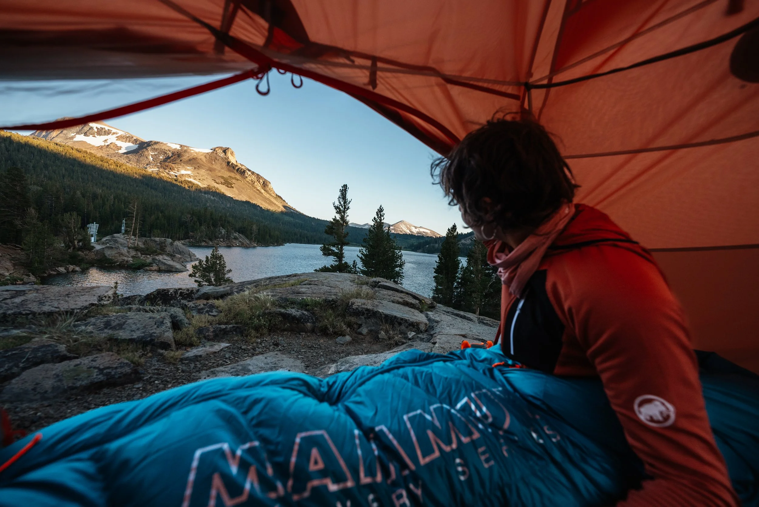 Person inside a tent looking out at a mountain lake landscape with trees and snow-capped peaks.