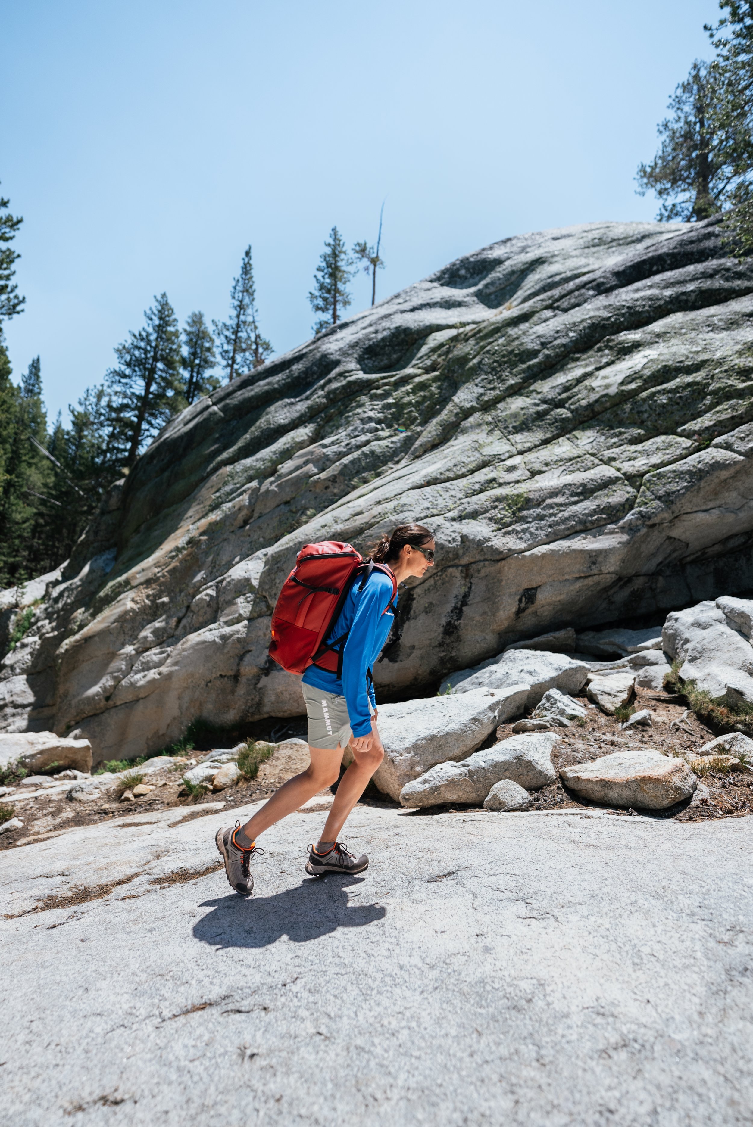 A woman hiking in a rocky outdoor area, carrying a red backpack, wearing a blue jacket, gray shorts, and hiking shoes, with tall trees and large rock formations in the background.