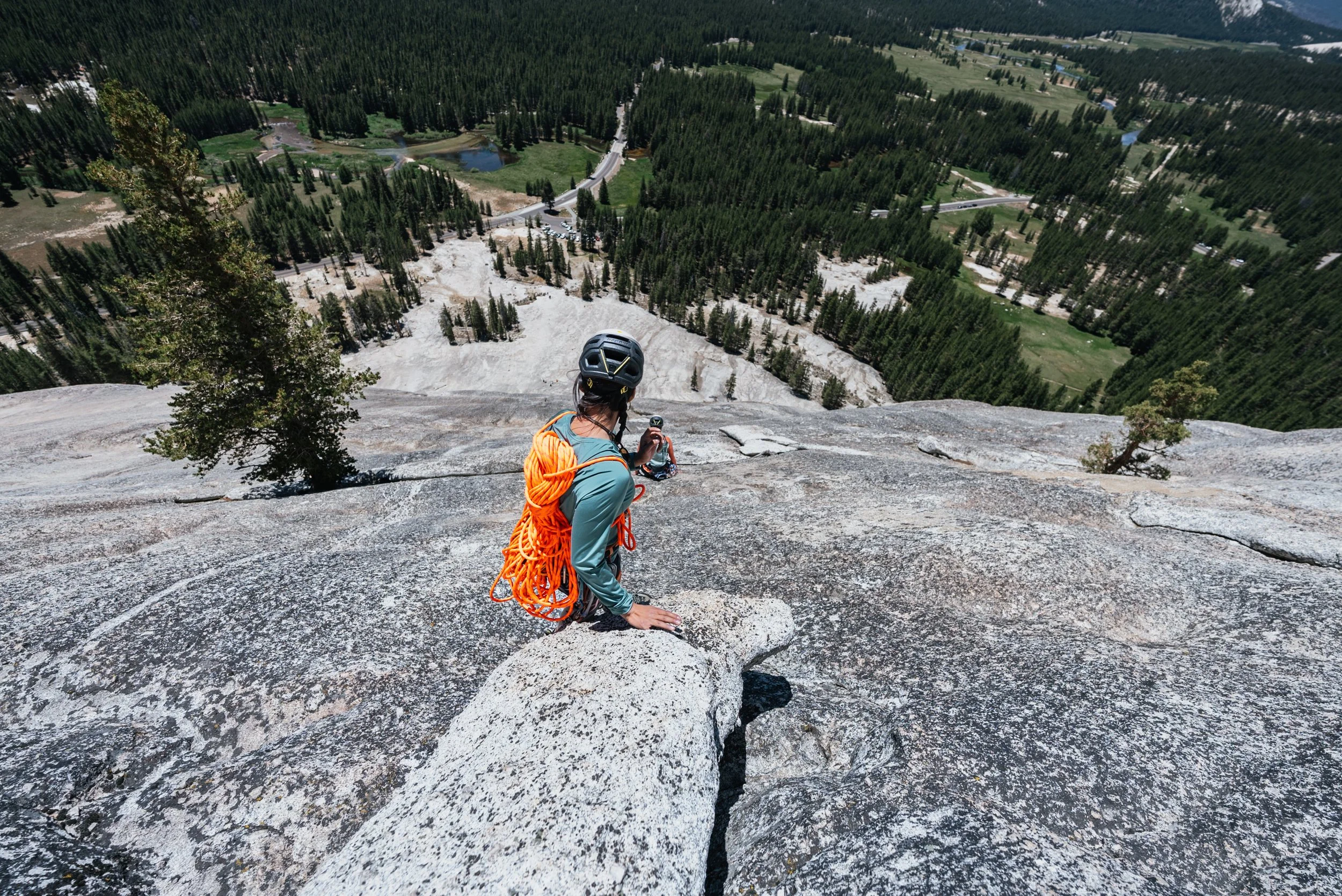A rock climber or mountaineer sitting on a large granite rock face overlooking a forested landscape with roads, lakes, and green fields.