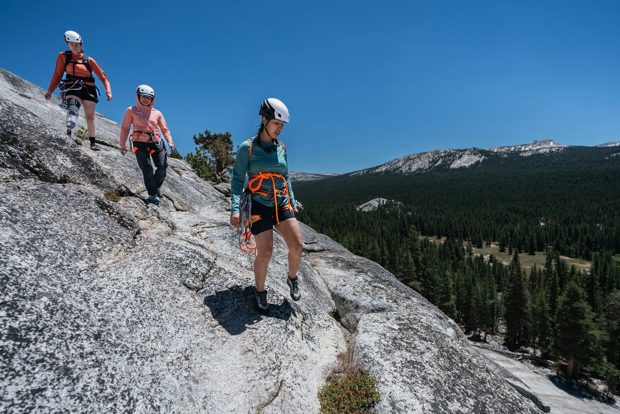 Four women rock climbing on a steep granite slope in a mountainous forested landscape under a clear blue sky, wearing helmets and climbing gear.