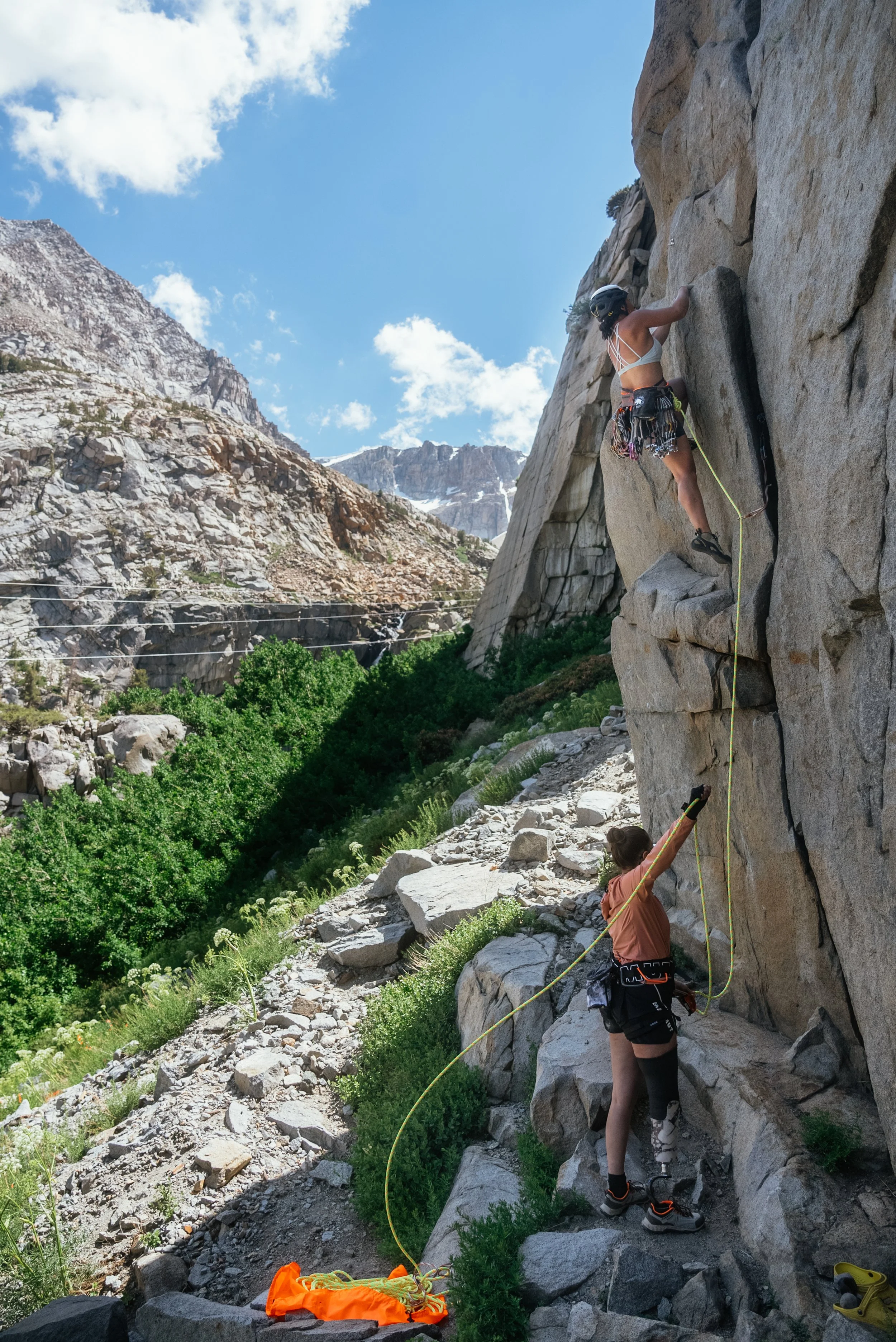 A woman rock climbing on a steep outdoor cliff with a guide below, against a backdrop of mountainous terrain and a partly cloudy sky.
