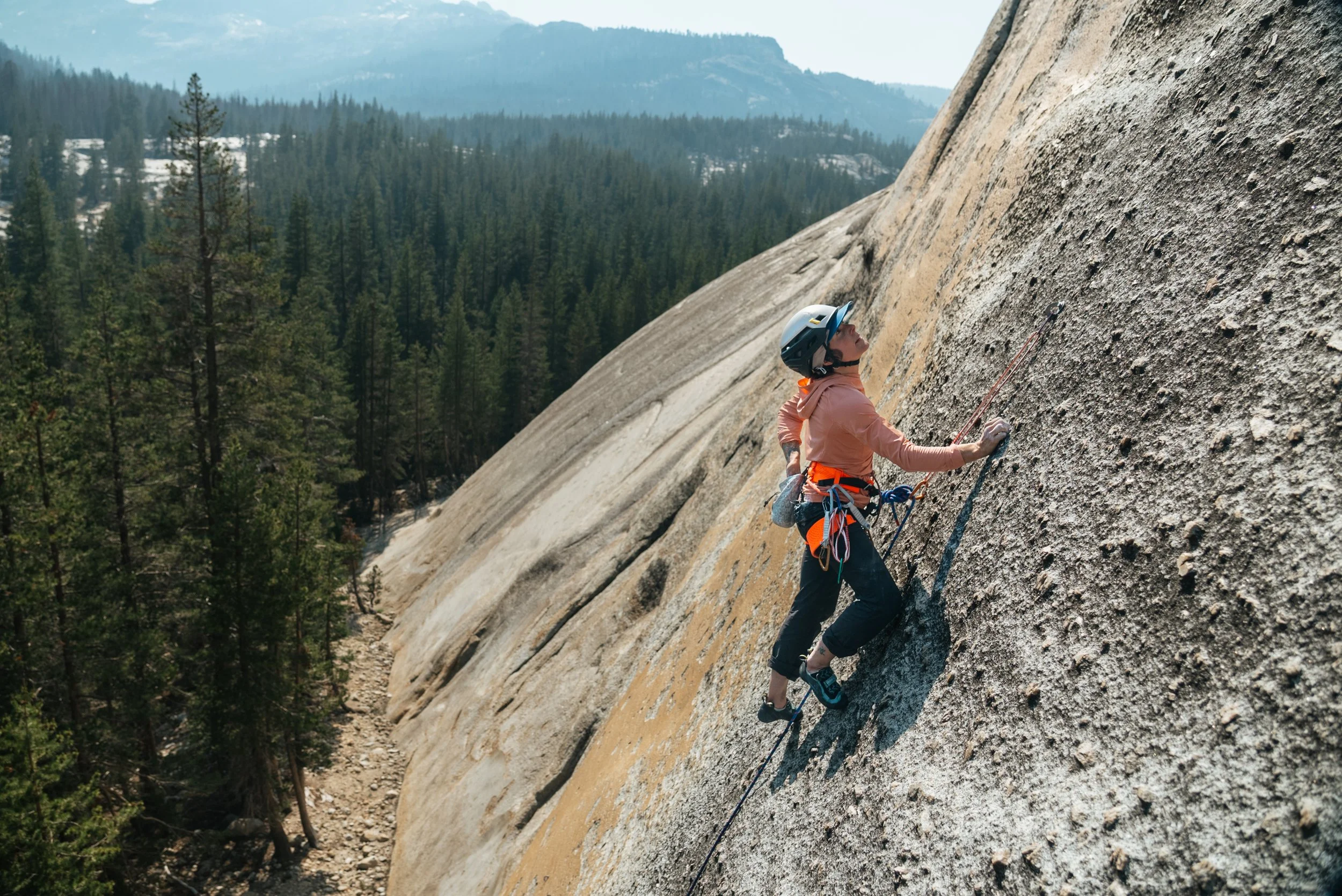 A person wearing a helmet and climbing gear is rock climbing on a steep granite wall in a forested mountain area.