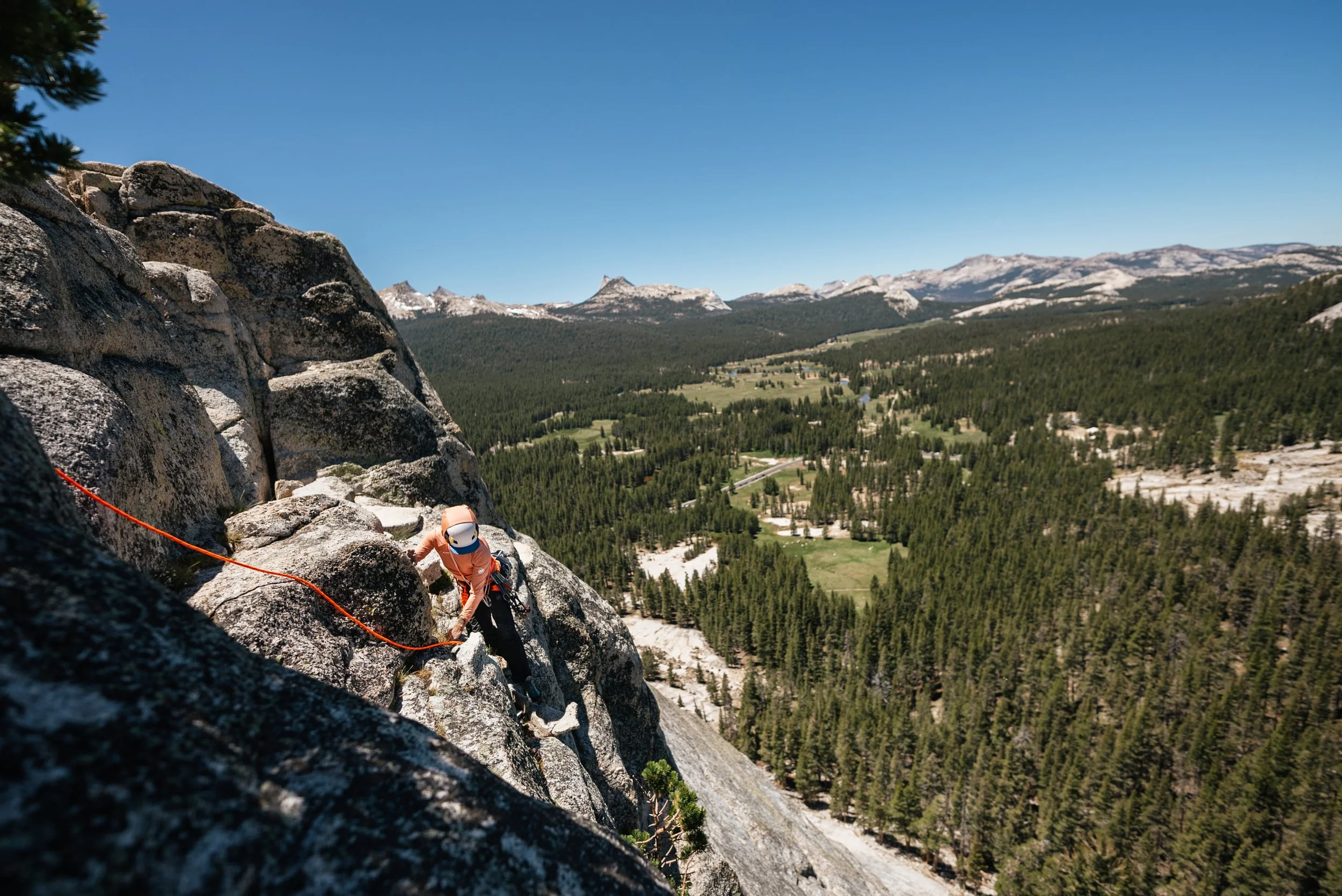 A person rock climbing on a steep granite cliff with a safety rope, overlooking a forested valley and mountain range under a clear blue sky.