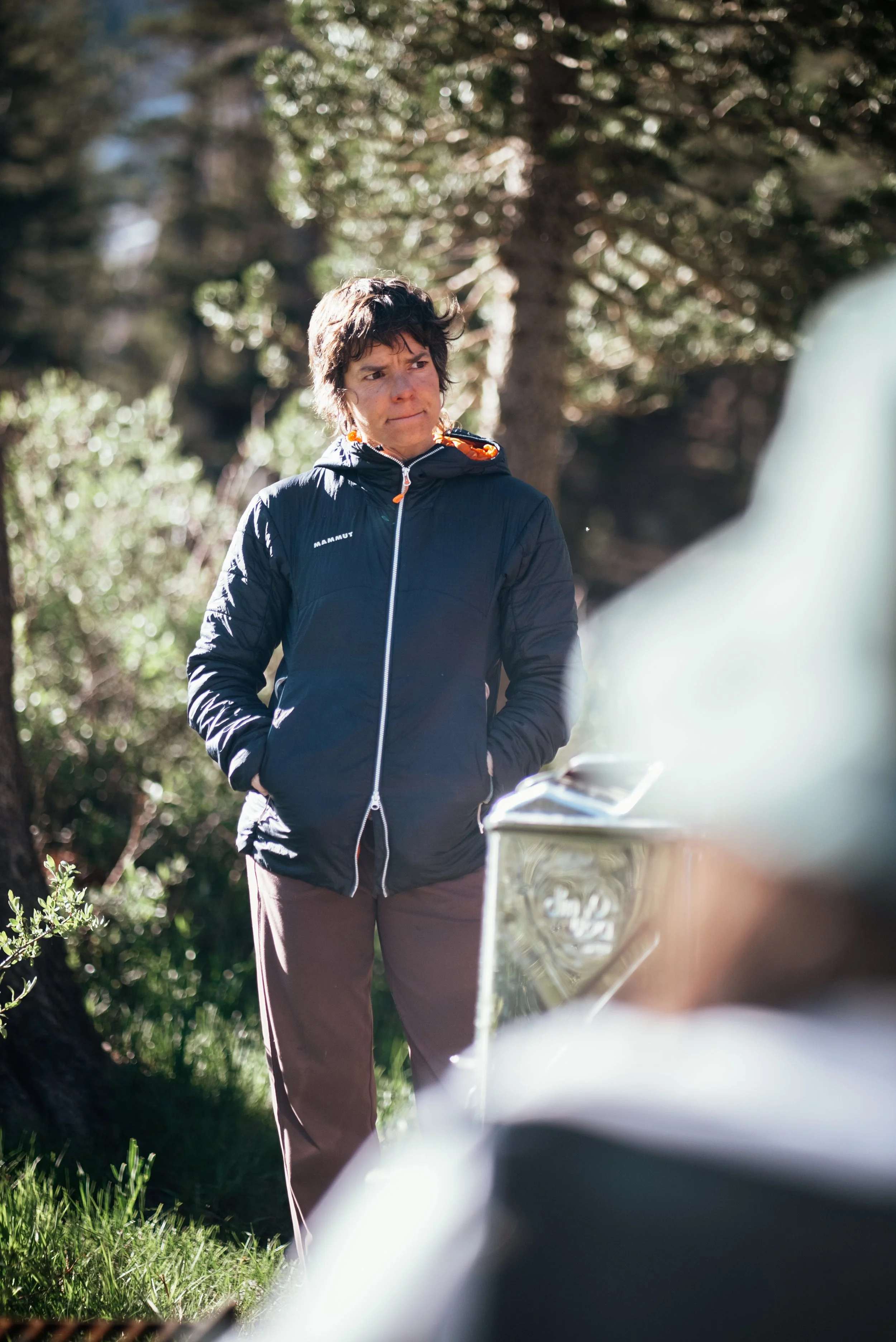 A woman with short dark hair wearing a black jacket standing outdoors among trees, looking to the side with a thoughtful expression.