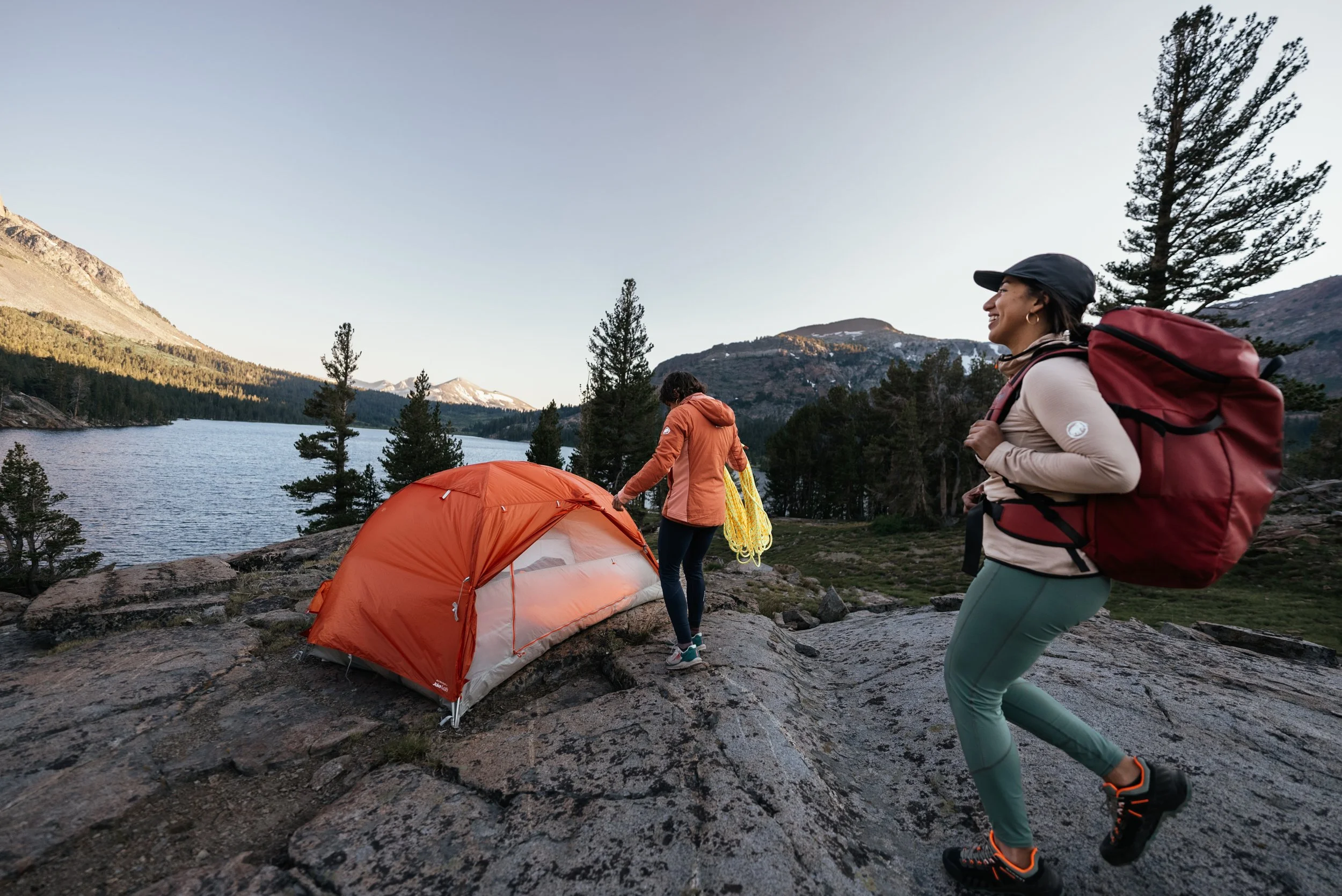 Two women camping near a lake with mountains in the background. One woman is setting up or checking an orange tent, while the other woman, carrying a large red backpack, is smiling and walking on rocky terrain.