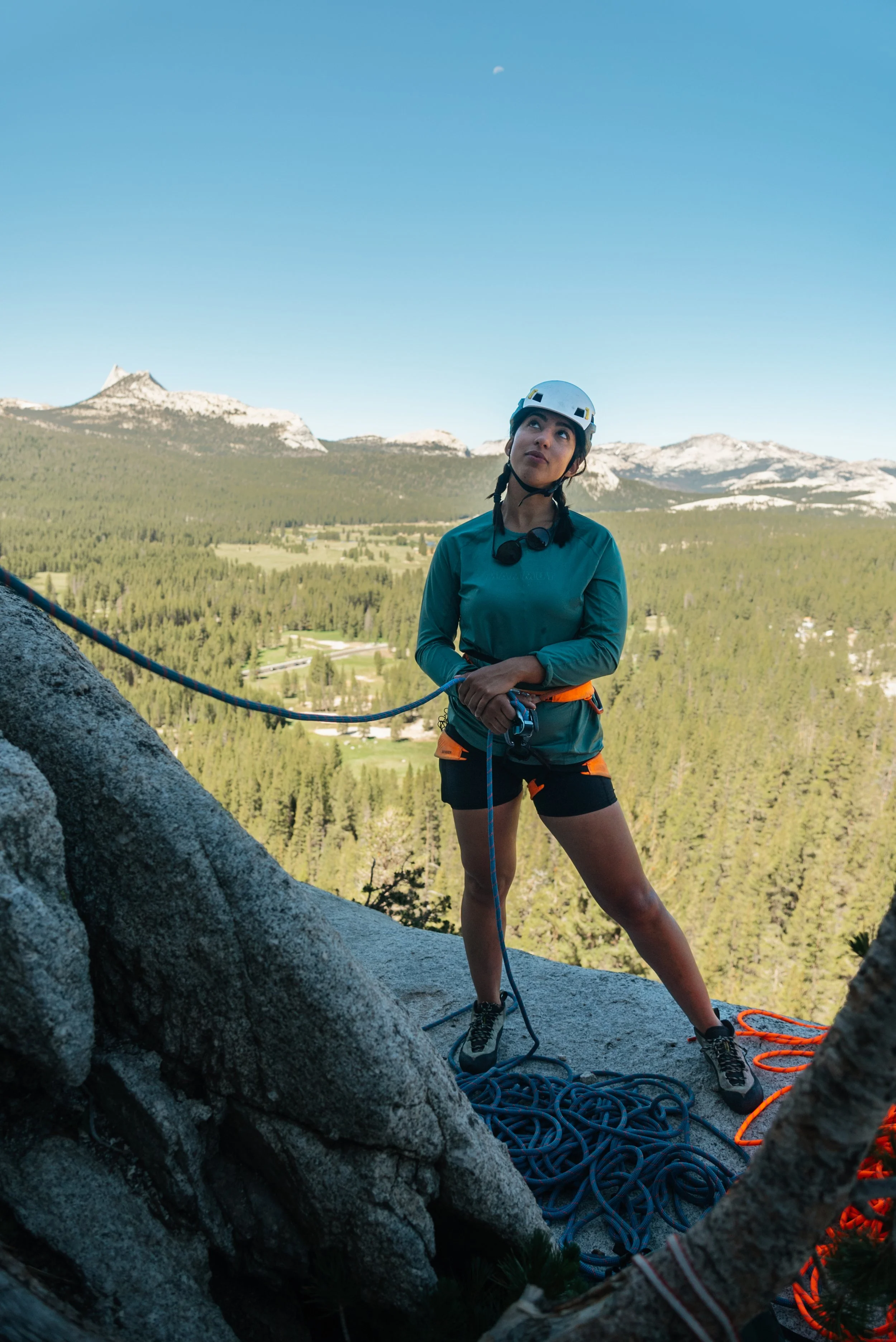 A woman wearing a helmet and climbing gear stands on a rock ledge with a scenic mountain and forest landscape in the background.
