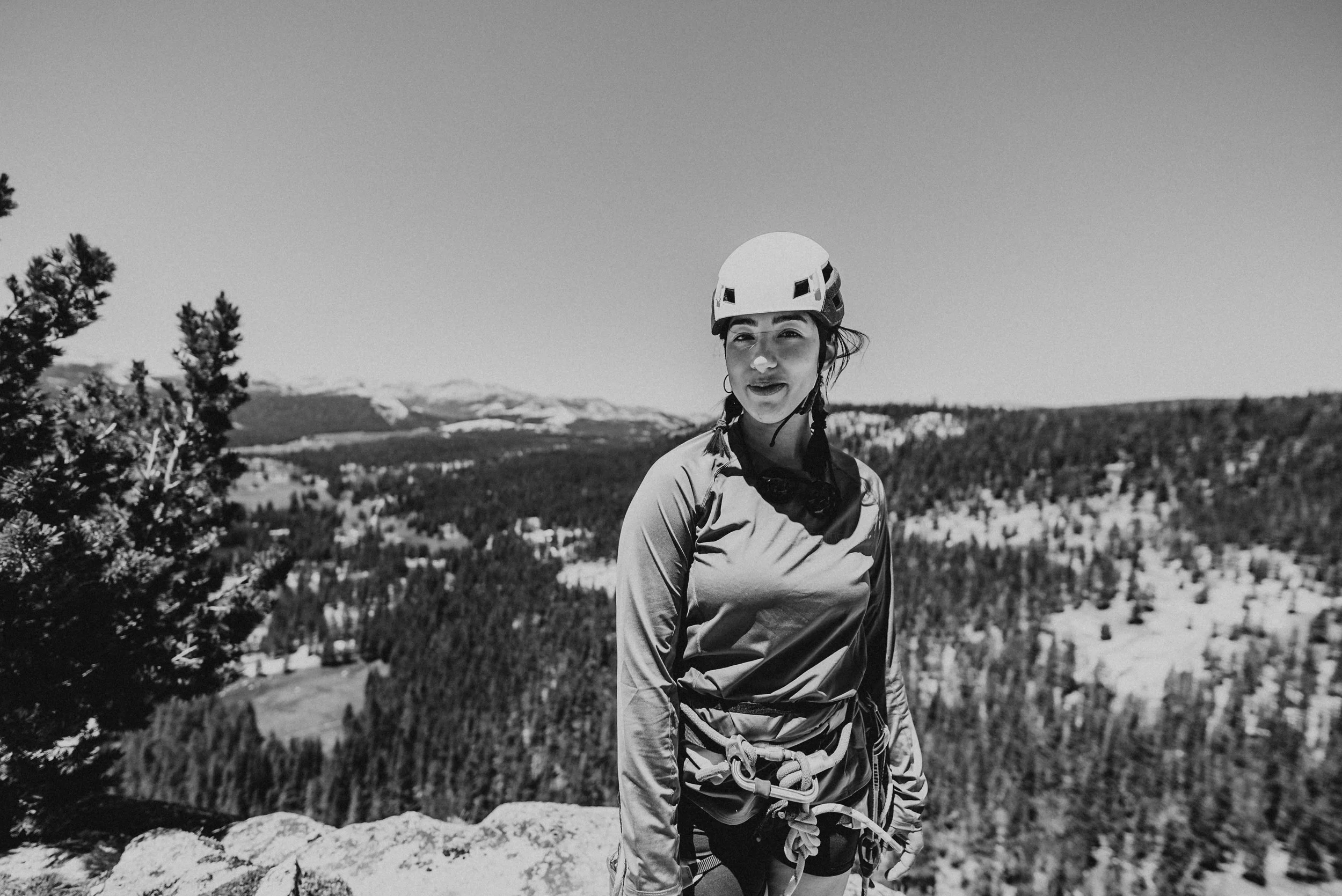 A woman wearing hiking gear and a helmet stands on a rocky ledge with a mountainous landscape in the background.