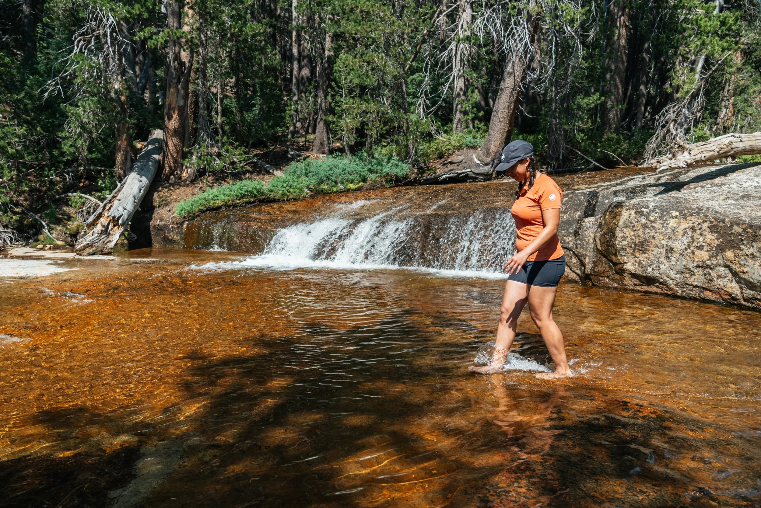 A woman in an orange shirt, black shorts, and a black cap standing in shallow water near a small waterfall in a forested area.