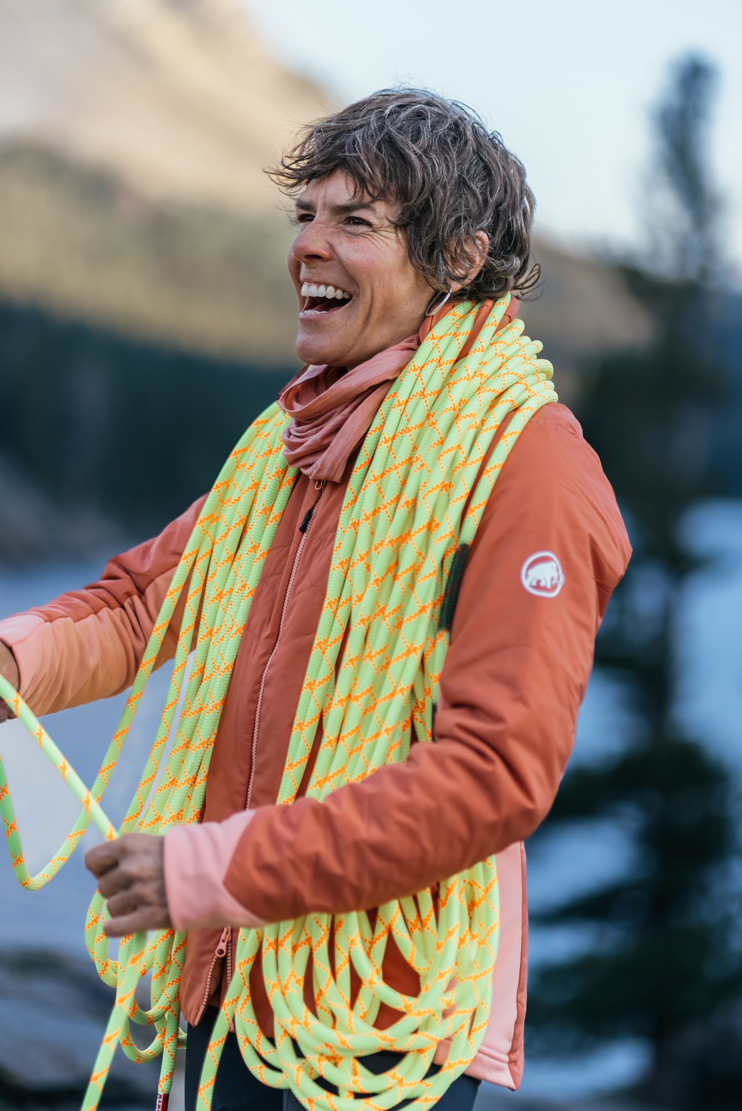 Smiling woman outdoors with a yellow climbing rope draped over her shoulders, wearing an orange jacket and pink scarf, with blurred trees and water in the background.