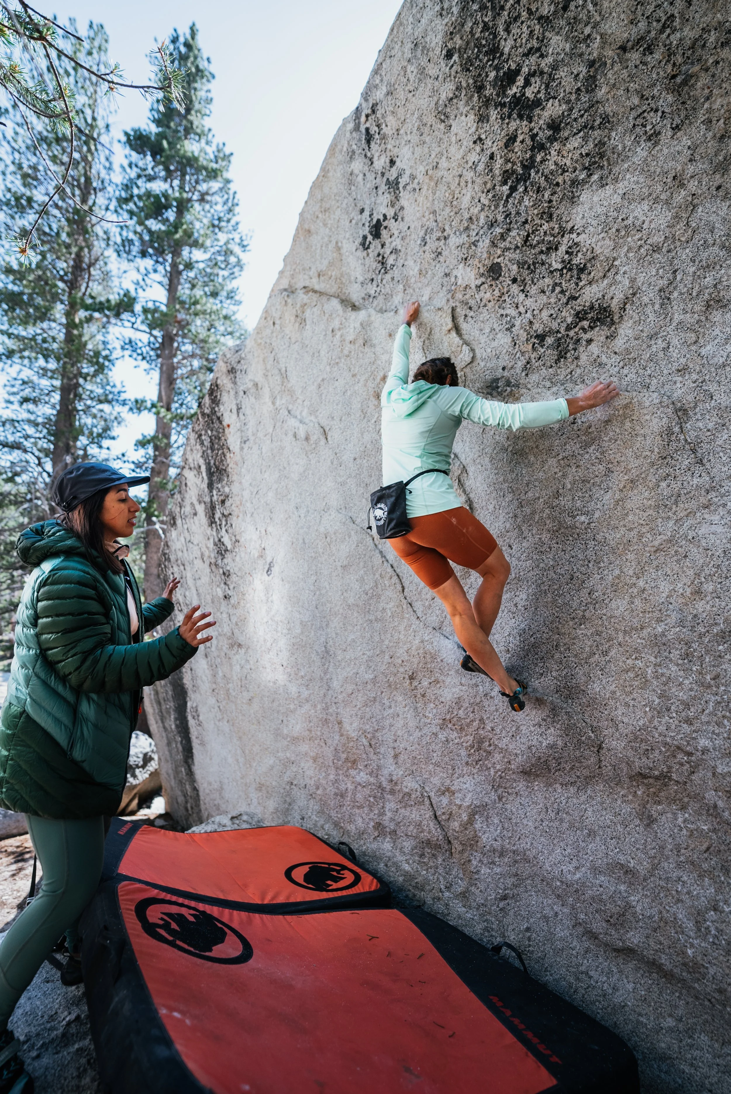 A woman bouldering on a large granite rock wall outdoors, with a spotter standing nearby ready to assist, and a crash pad at the base for safety, surrounded by trees.
