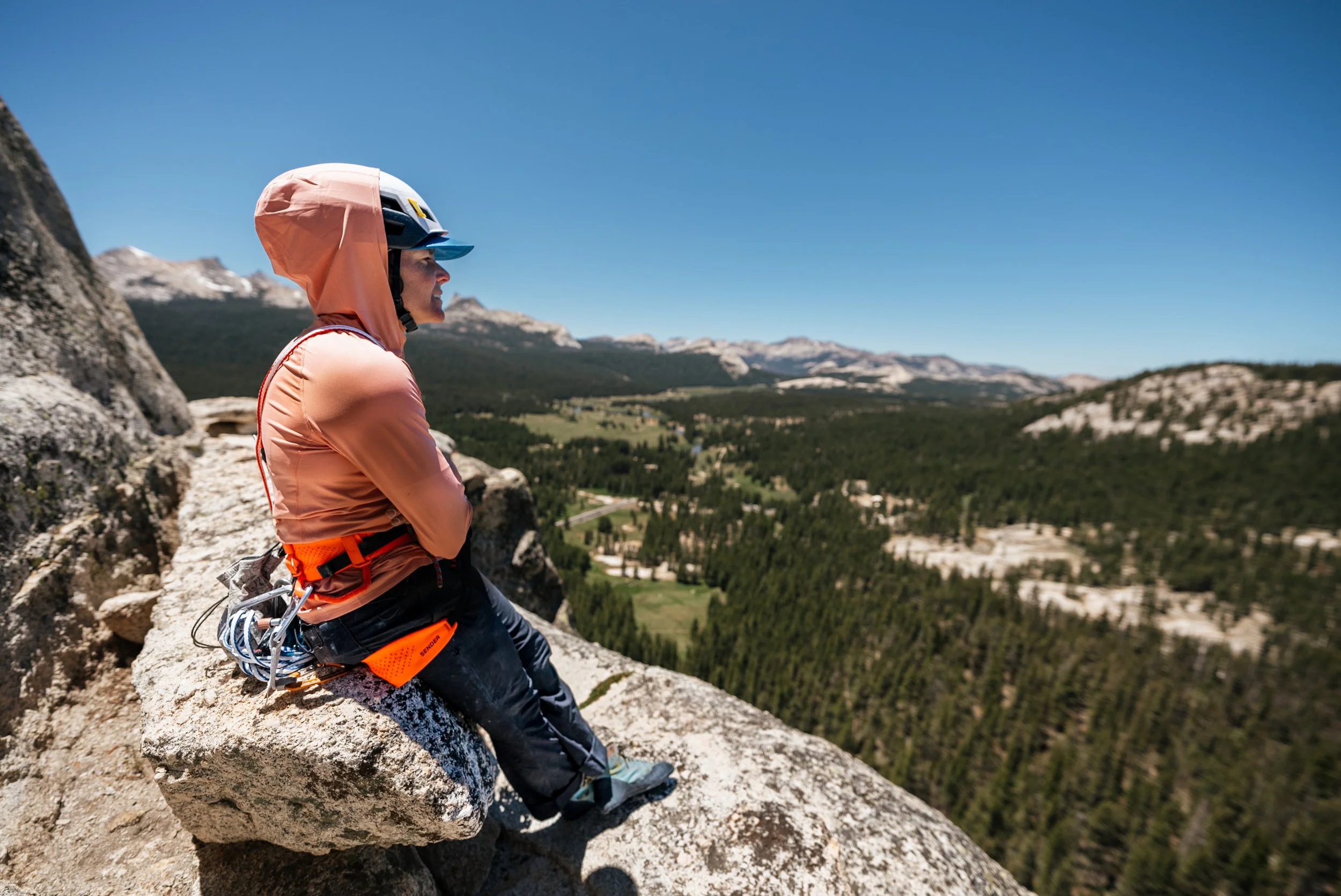 A woman sitting on a rock ledge overlooking a mountainous landscape with forests under a clear blue sky, wearing a pink jacket, helmet, and climbing gear.
