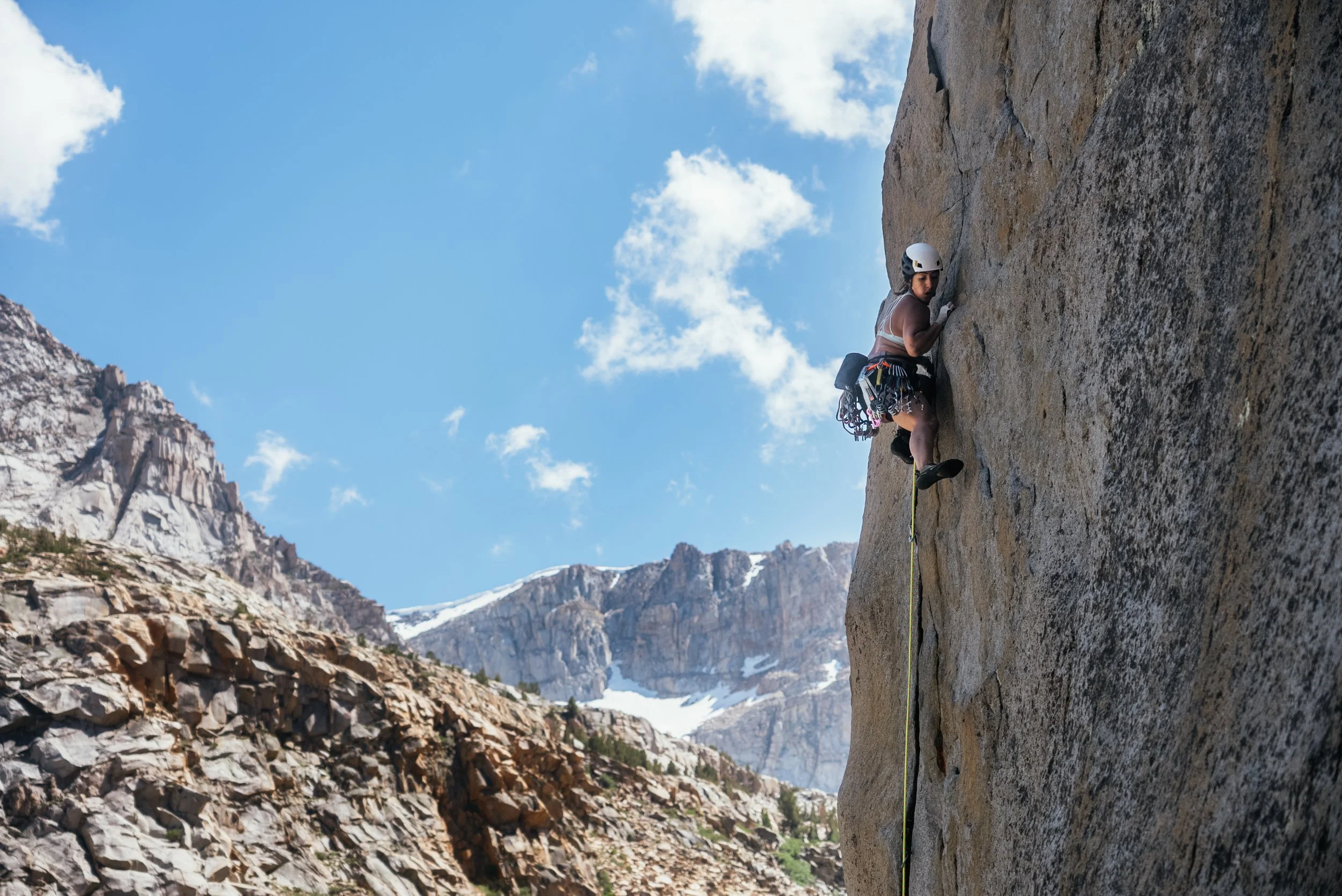 Woman rock climbing on a vertical cliff face against a mountain landscape and blue sky.