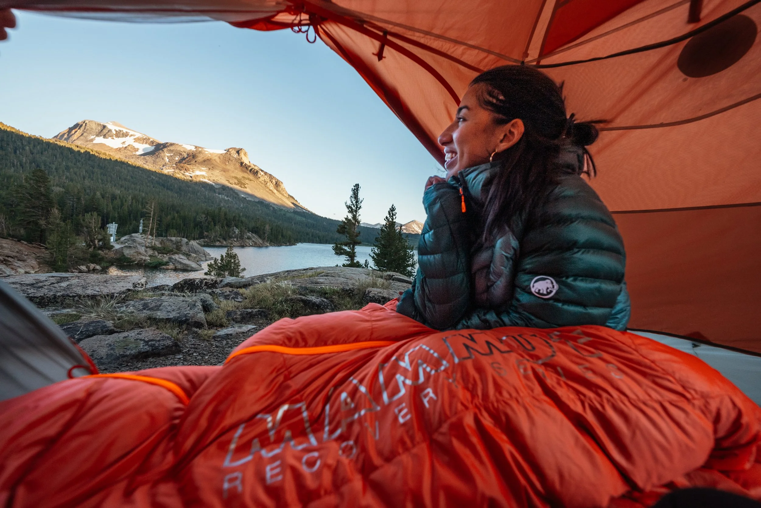 A woman sitting inside a tent outdoors, smiling and looking towards a scenic lake with mountains and trees in the background.