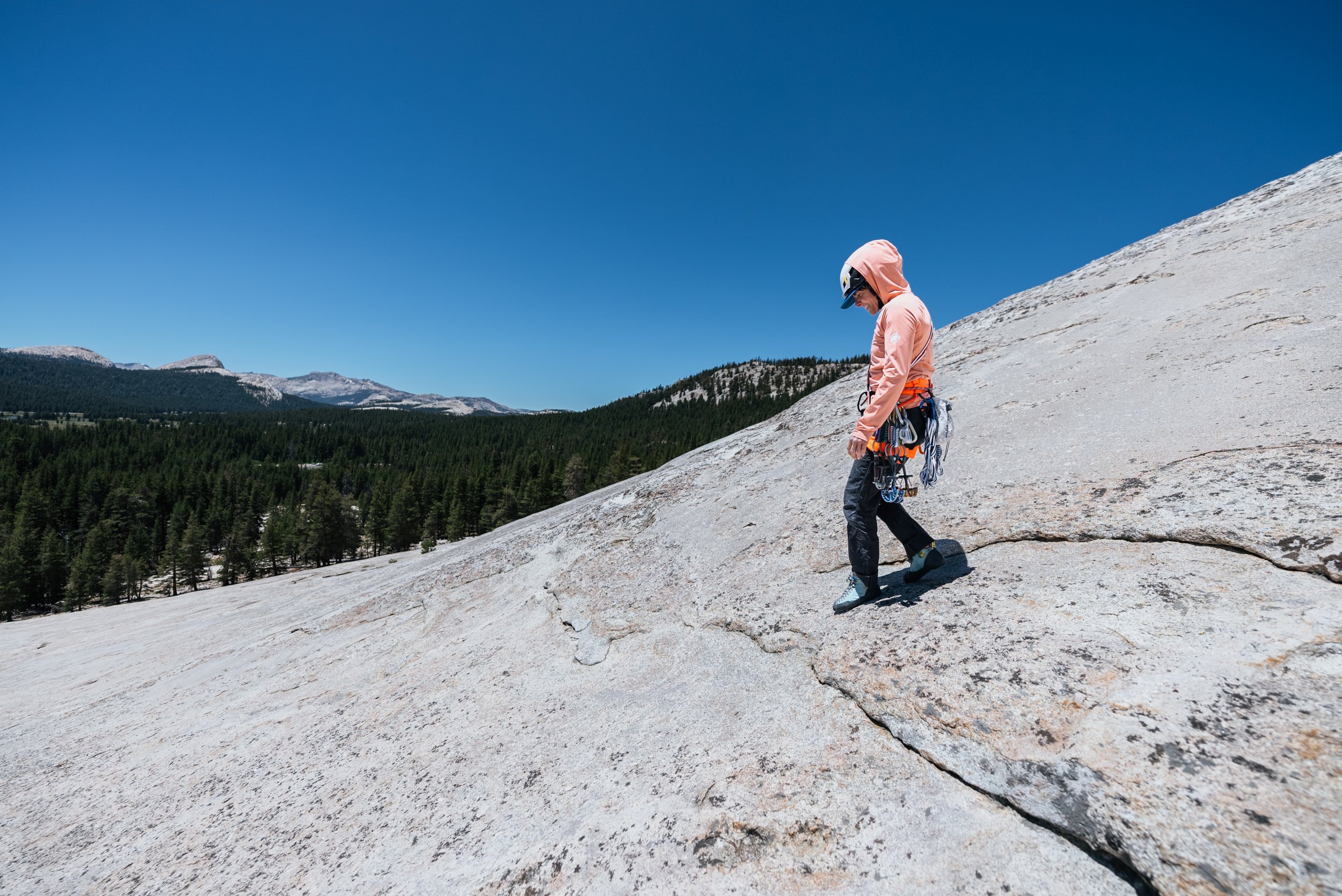 A person dressed in pink outdoor gear with climbing equipment is rock climbing on a large smooth granite surface under a clear blue sky, with a dense forest and mountain range in the background.