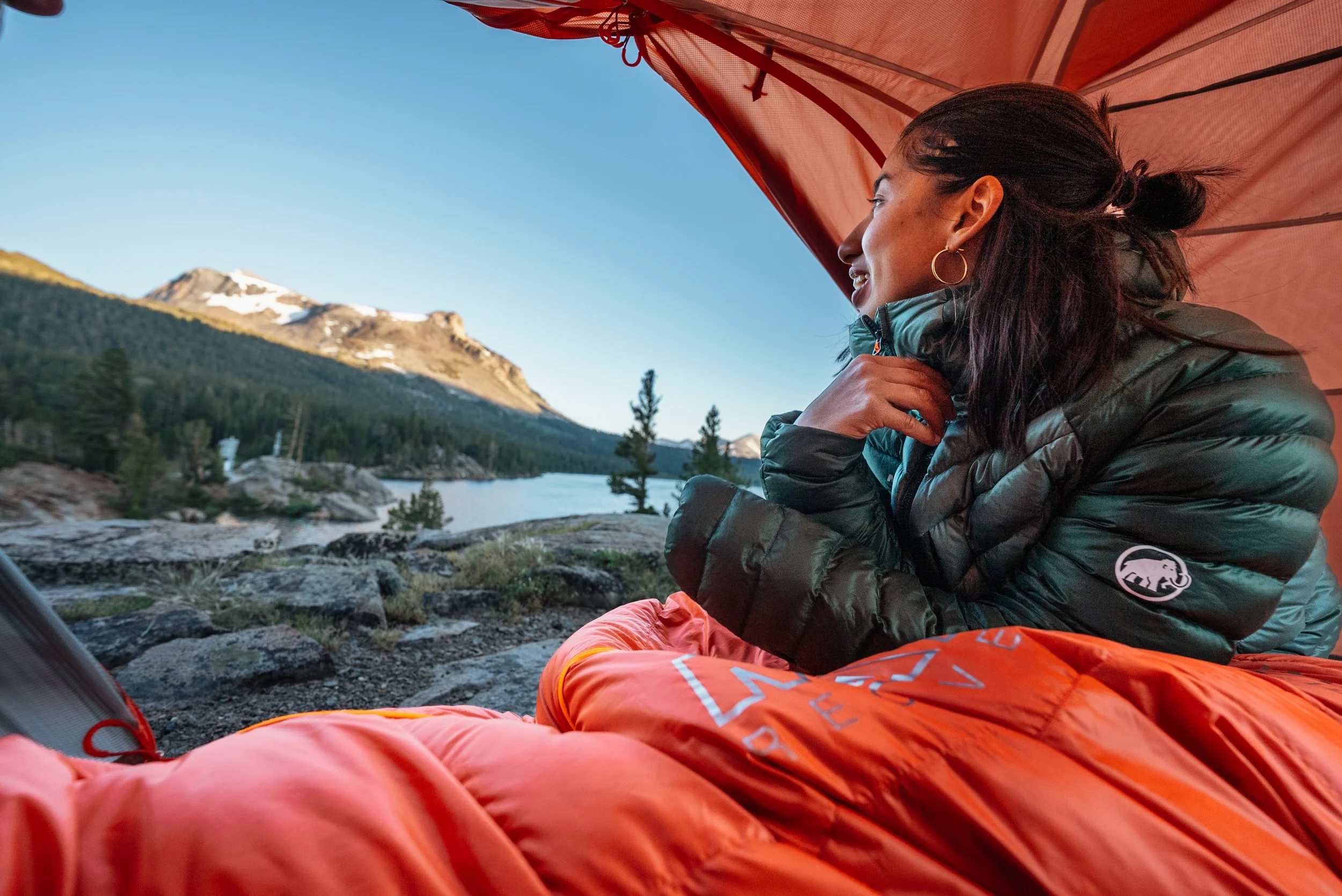 A woman sitting inside a tent looks out at a mountain landscape with snow-capped peaks, trees, and a lake, during daylight. She wears a green puffer jacket, hoop earrings, and has her hand on her chin, resting on her knee.