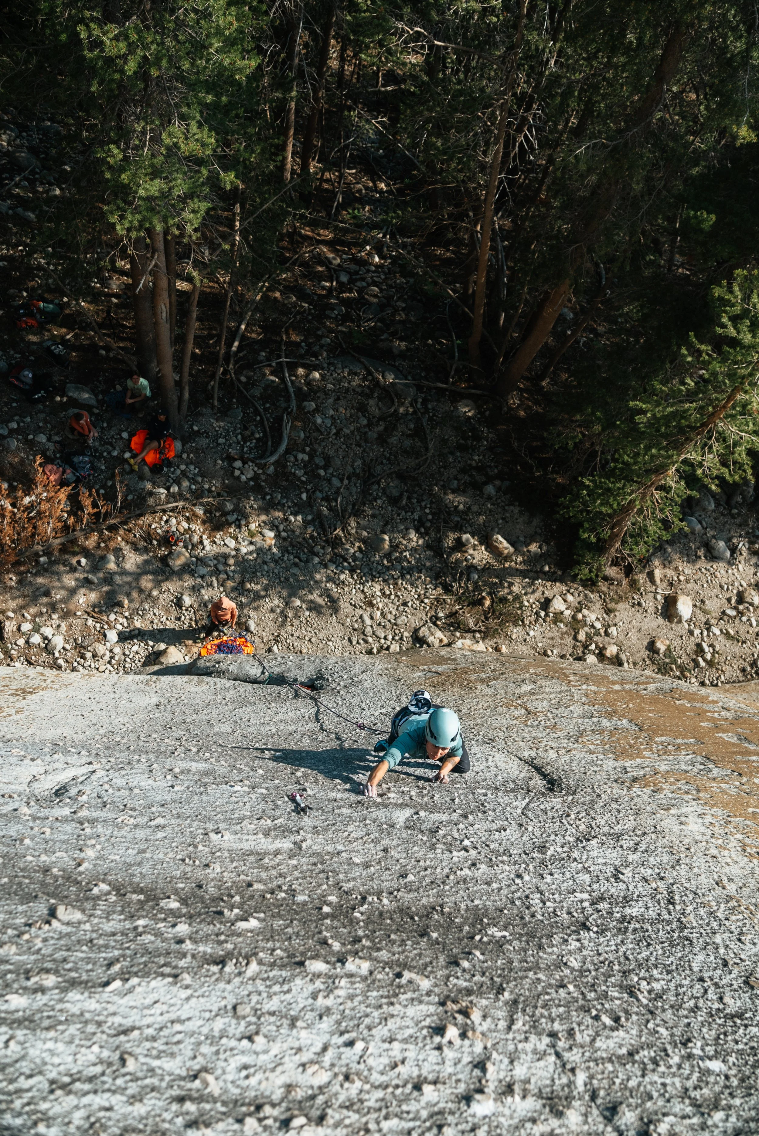 A person rock climbing on a steep, gray rock face with trees and other climbers below.