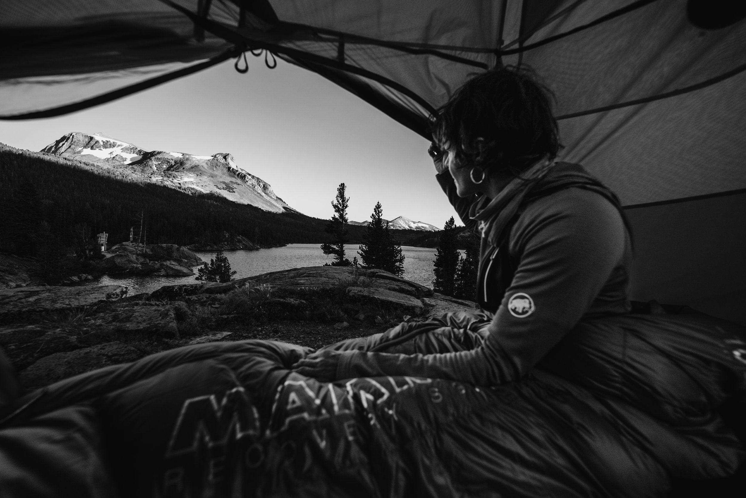 A woman sitting inside a tent, looking out at a mountain lake with snow-capped peaks and pine trees in the distance.