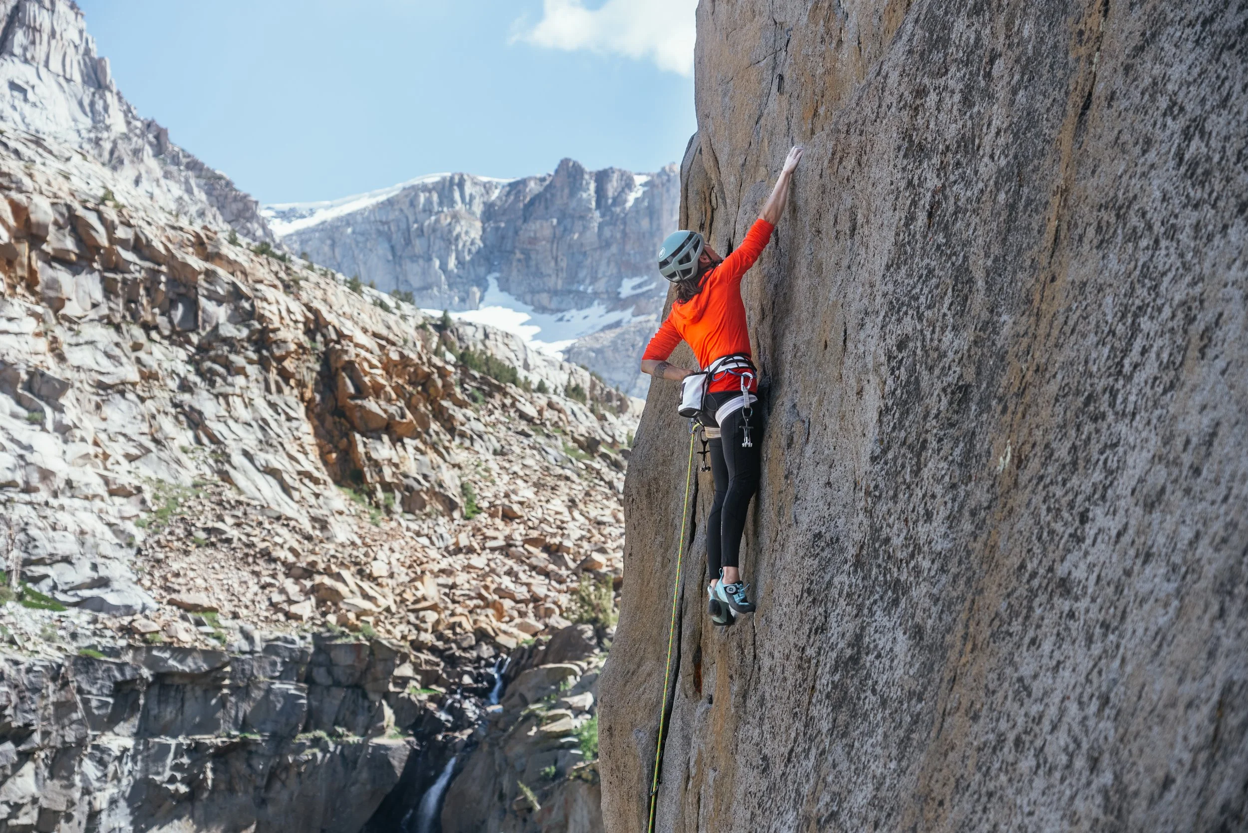 A woman rock climbing on a large steep granite wall in a mountainous area with snow-capped peaks in the background.